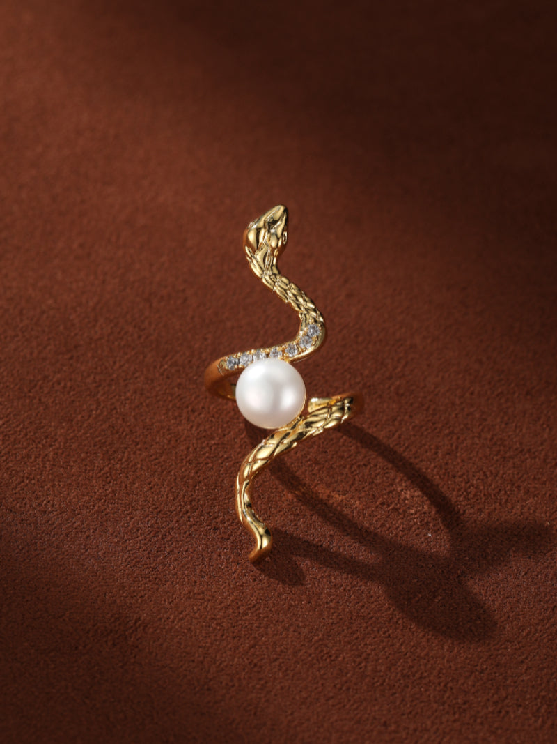 Close-up of a gold-tone snake cuff ring with a white freshwater pearl, set against a textured brown background. Tiny clear stones accent the snake's body.