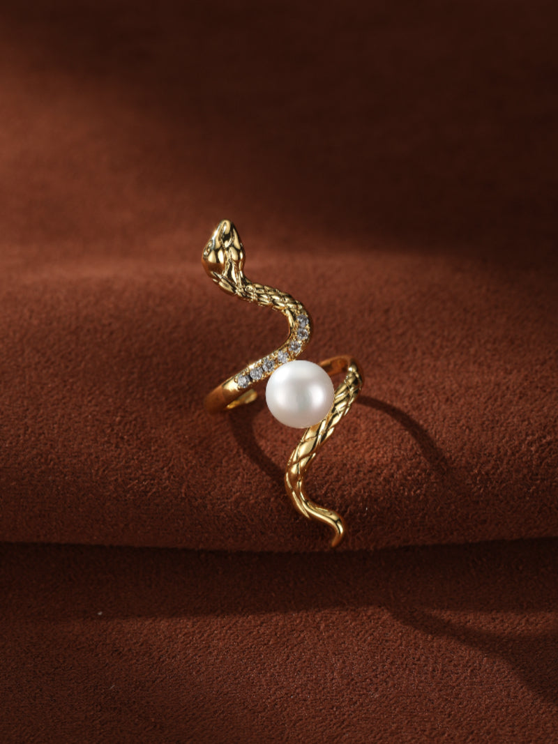 A close-up product shot of the Eternal Coil Freshwater Pearl Snake Cuff Ring, featuring a detailed gold snake design with small clear accent stones, elegantly coiled around a lustrous white freshwater pearl. The ring rests on a textured brown surface. @pearl