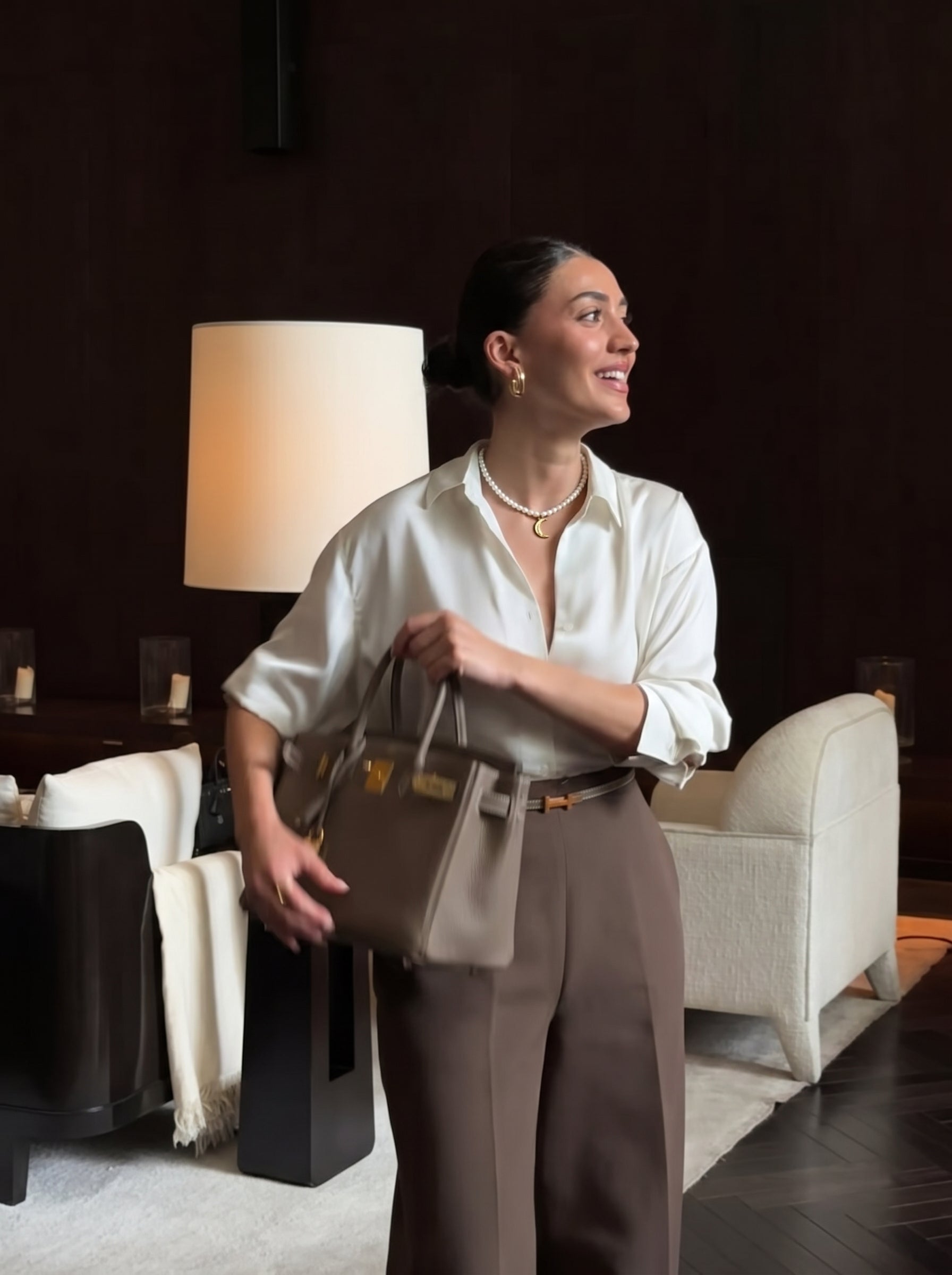 A woman wearing a white blouse, brown pants, and a freshwater pearl choker necklace with a golden moon pendant smiles and holds a handbag. @lifestyle #lifestyle