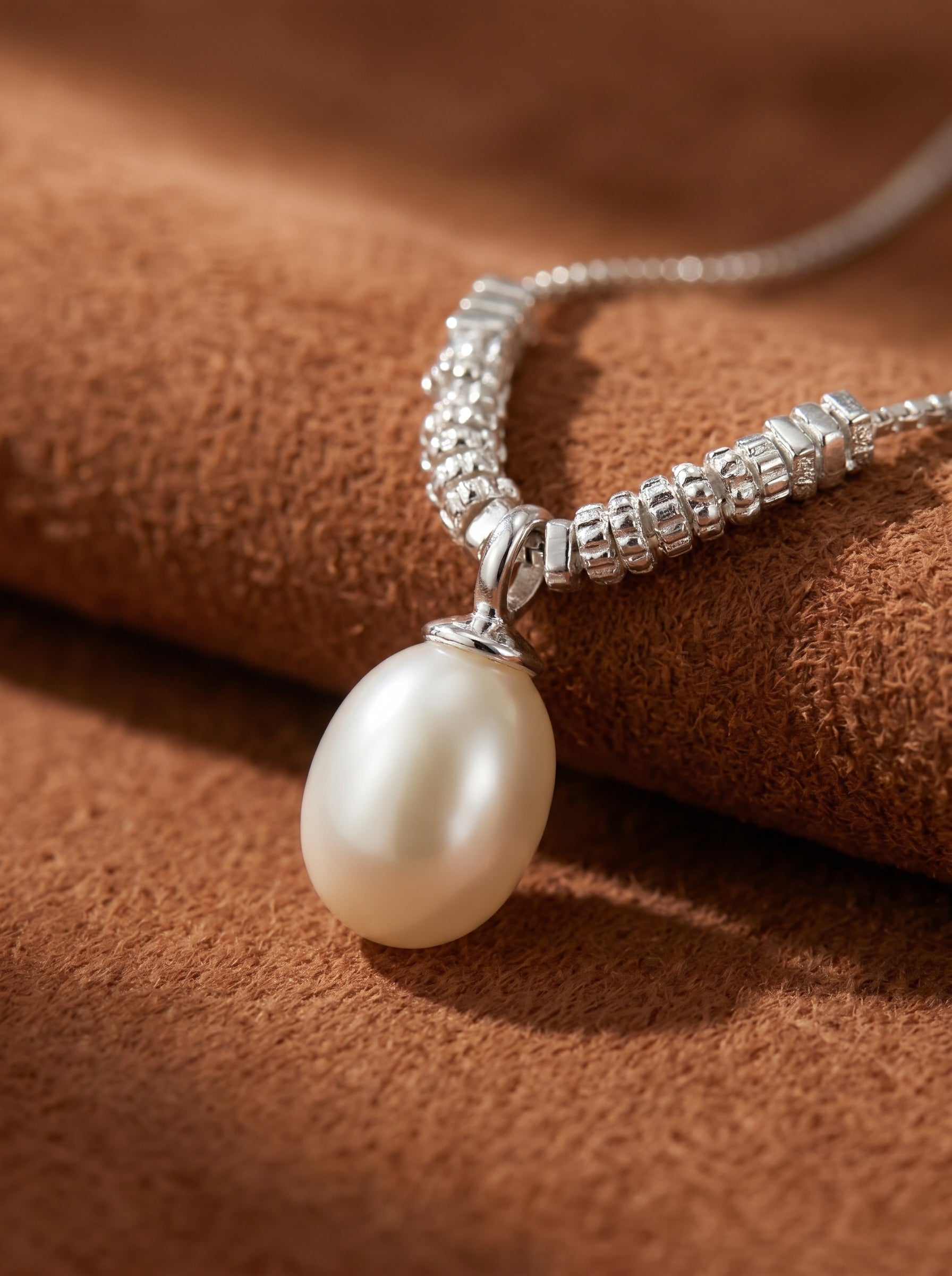 A close-up of a silver-toned necklace with a textured chain and a single, large, oval-shaped white freshwater pearl drop. The pearl has a beautiful luster and is suspended from the chain. The necklace is displayed on a textured brown background. @pearl
