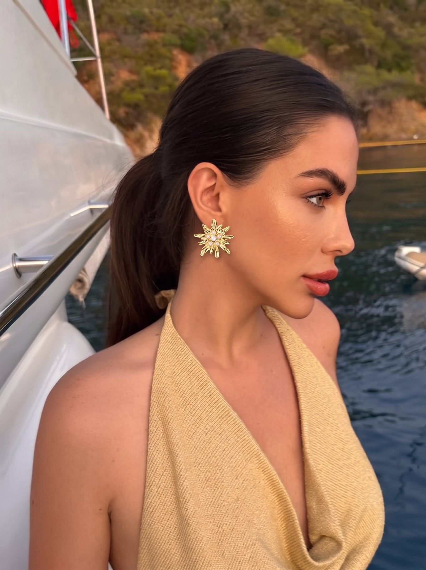 Model wearing gold-toned flower stud earrings with imitation pearls, on a yacht with the ocean in the background. @lifestyle