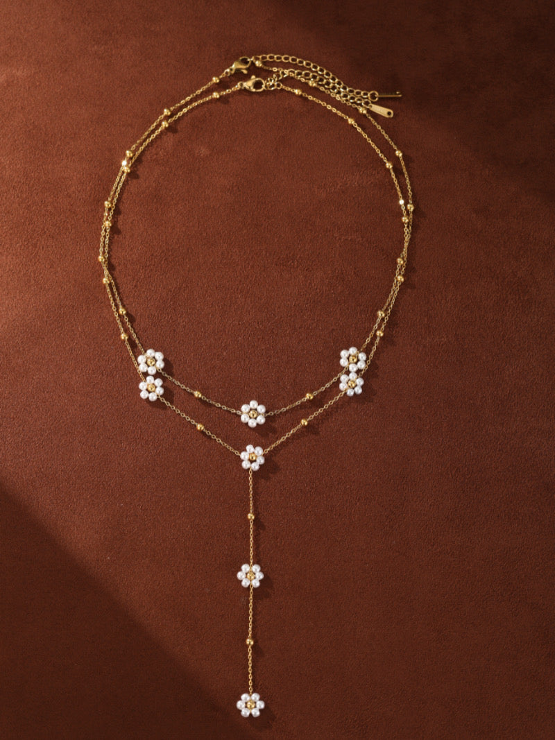 A delicate gold-toned necklace with two layered chains, featuring small white pearl flower charms and tiny gold beads. The necklace drapes in a Y-shape, with flowers cascading down the front.