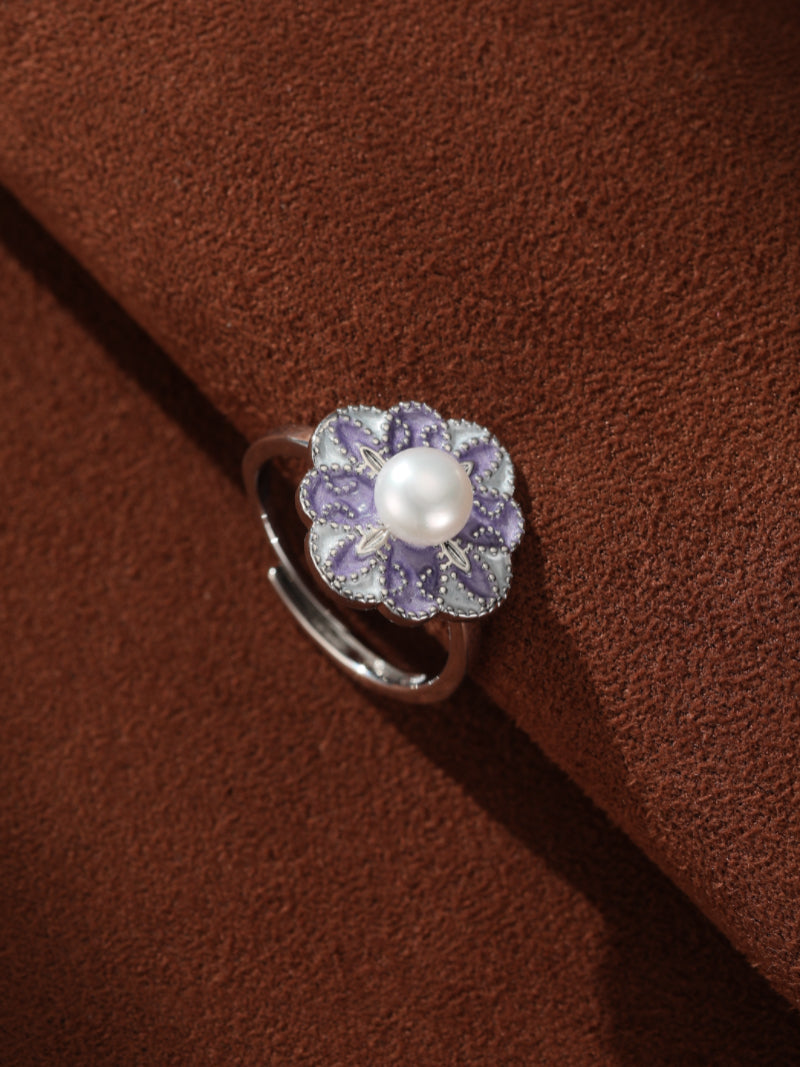A silver adjustable cuff ring featuring a purple enameled flower with a white freshwater pearl at its center, set against a brown textured background.