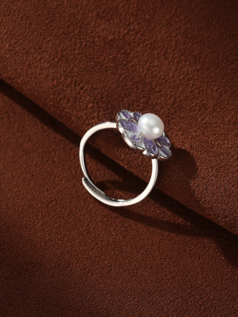 A silver cuff ring with a purple enameled flower motif cradling a white freshwater pearl, set against a brown textured background. #lifestyle