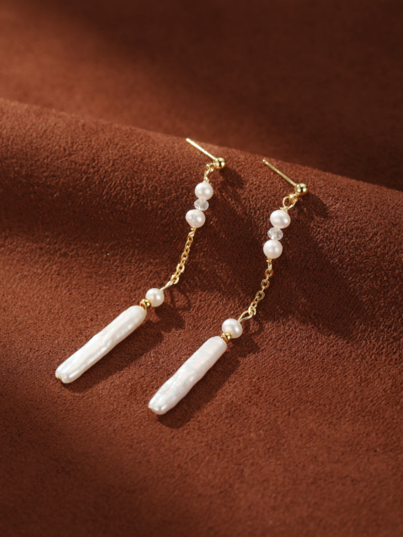 Flow - Freshwater Pearl Long Dangle Earrings featuring delicate white seed pearls and elongated white baroque pearls on a gold chain, set against a rich brown textured background.