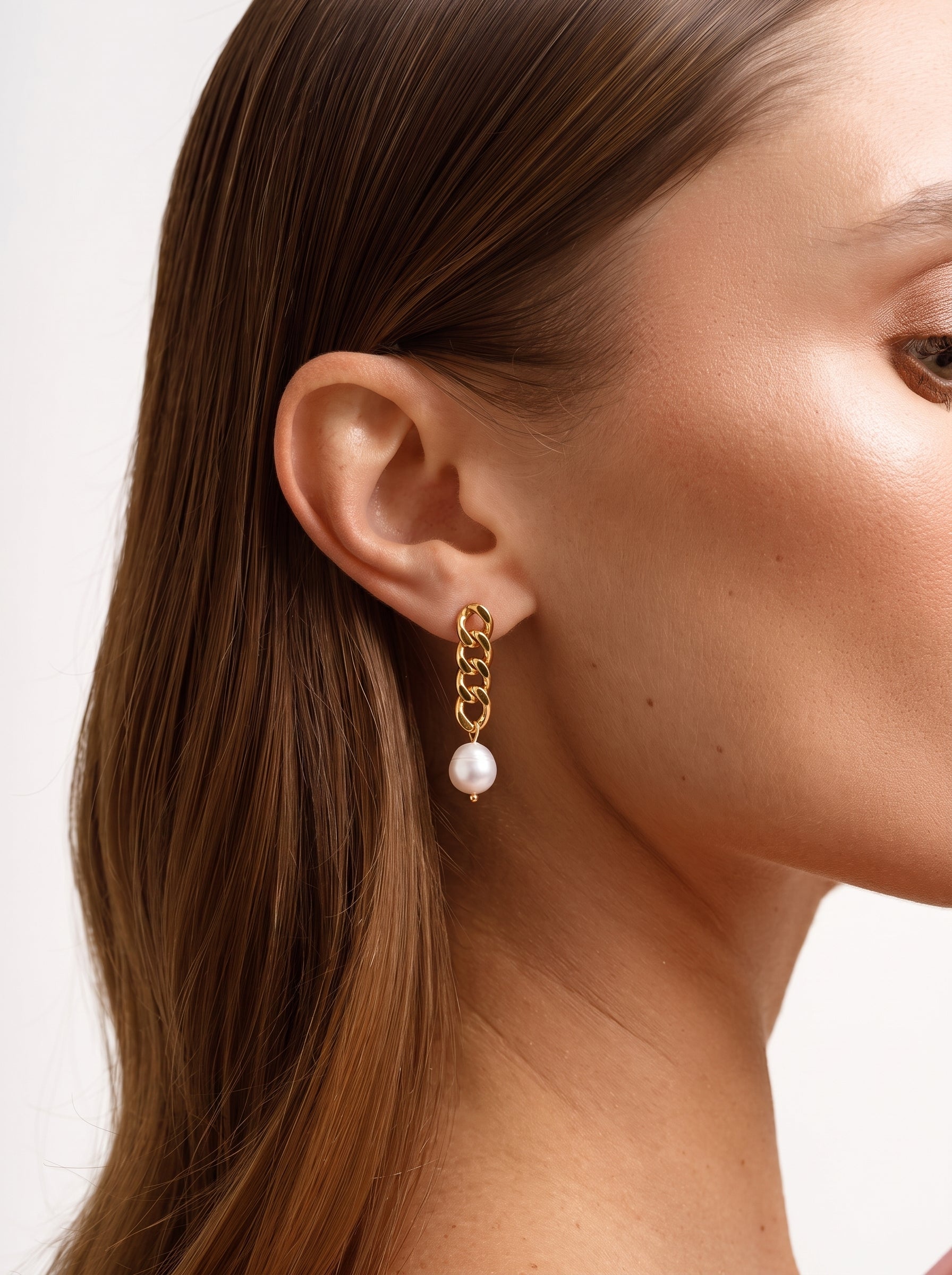 Close-up of a woman's ear wearing gold chain dangle earrings with a single white freshwater pearl at the end. @model