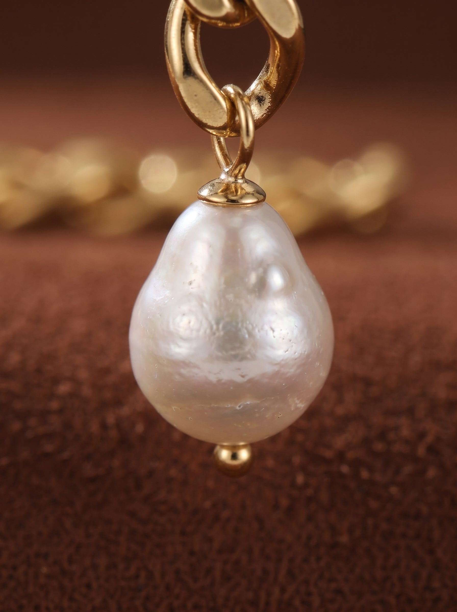 Close-up of a gold chain dangle earring featuring a lustrous white freshwater pearl with a seed shape and a small gold bead at the bottom. The earring is set against a textured brown background. @pearl