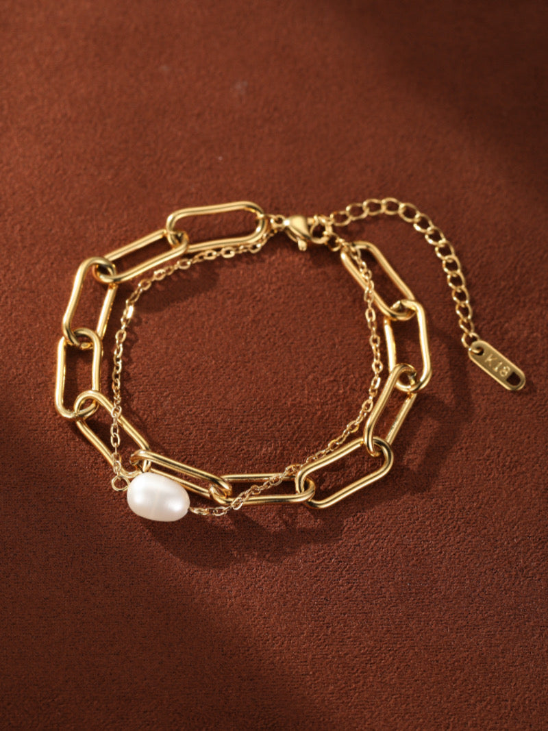 Gold-toned chunky chain bracelet with a delicate secondary chain, featuring a single white baroque freshwater pearl. The bracelet has an adjustable clasp and is shown against a brown textured background.
