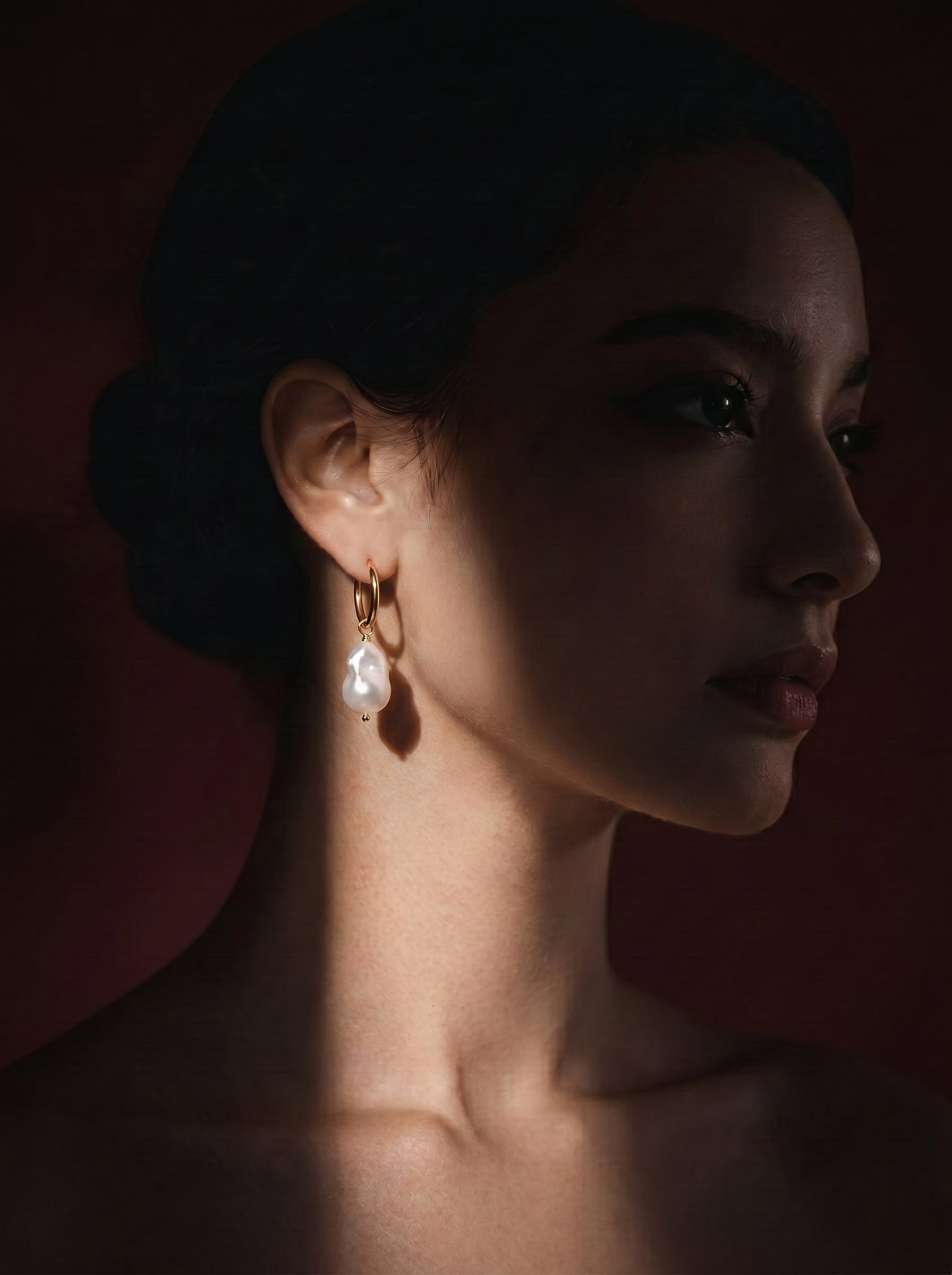 Close-up of a woman wearing a Forma freshwater pearl chunky hoop dangle earring. The earring features a gold hoop with a large, irregular white baroque pearl dangling from it. @model