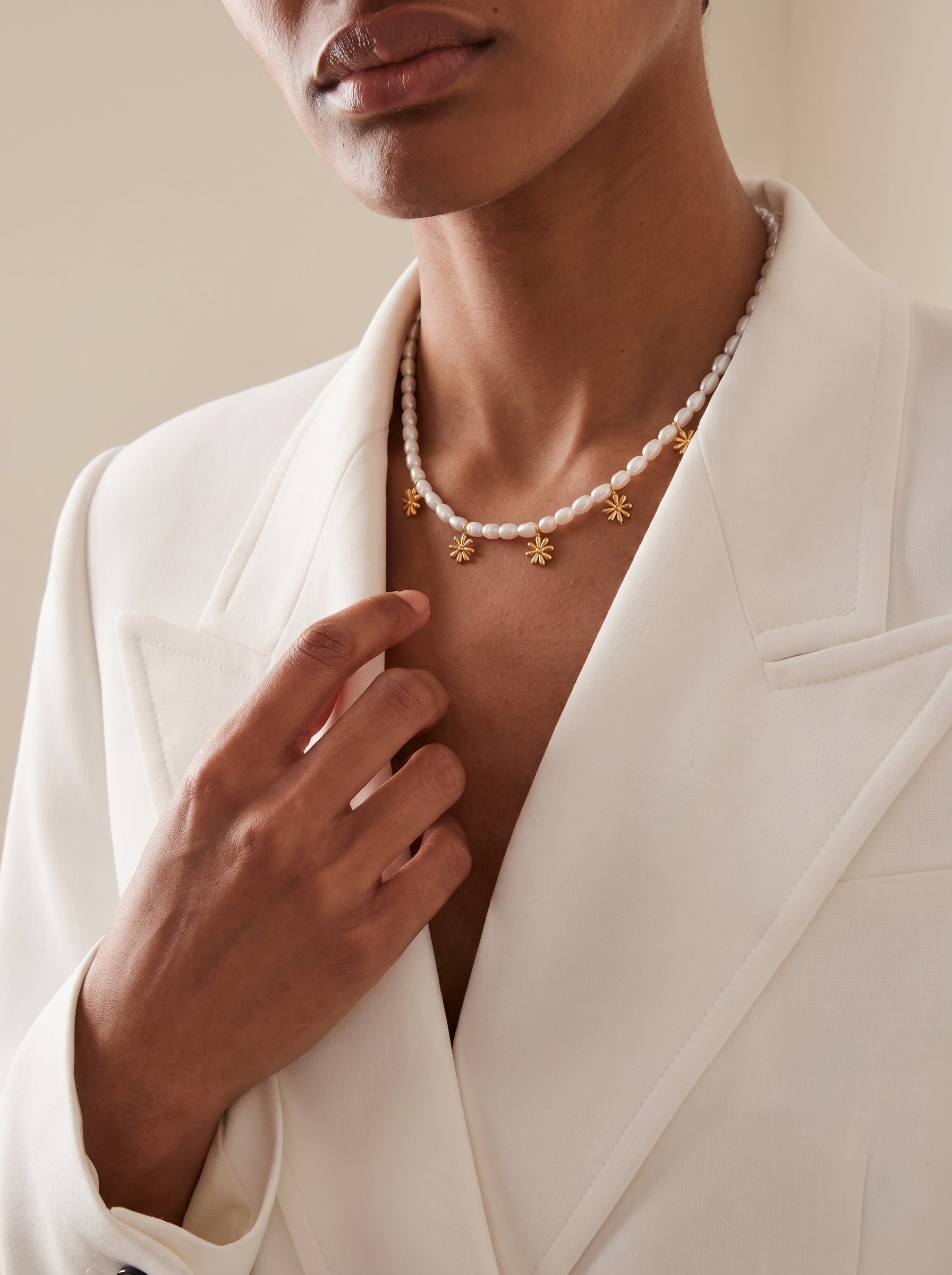A woman wearing a white blazer and a Gemma Strand choker necklace made of small white freshwater pearls with dangling gold flower charms. @model