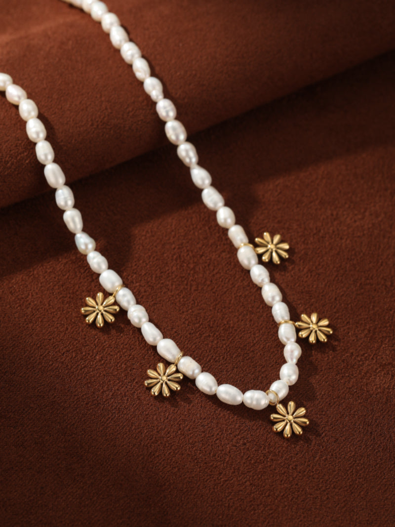Gemma Strand choker necklace featuring delicate white freshwater seed pearls and small gold flower charms, displayed against a textured brown background. @pearl