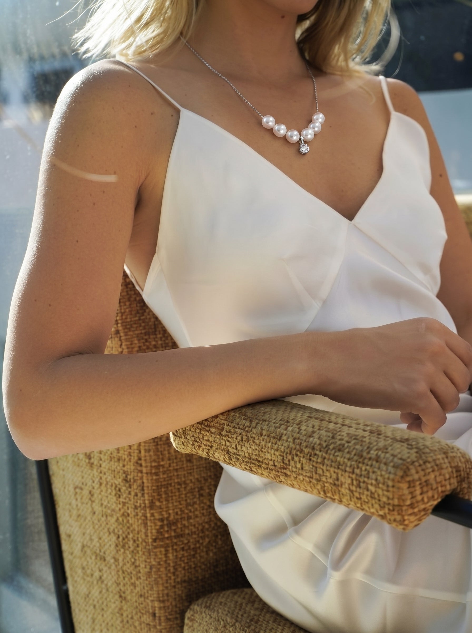 A woman in a white slip dress wears the Gleam Curve Freshwater Pearl Diamond Floating Necklace, featuring seven 6mm white pearls and a central diamond pendant, seated in a chair with her arm resting on the armrest. @lifestyle