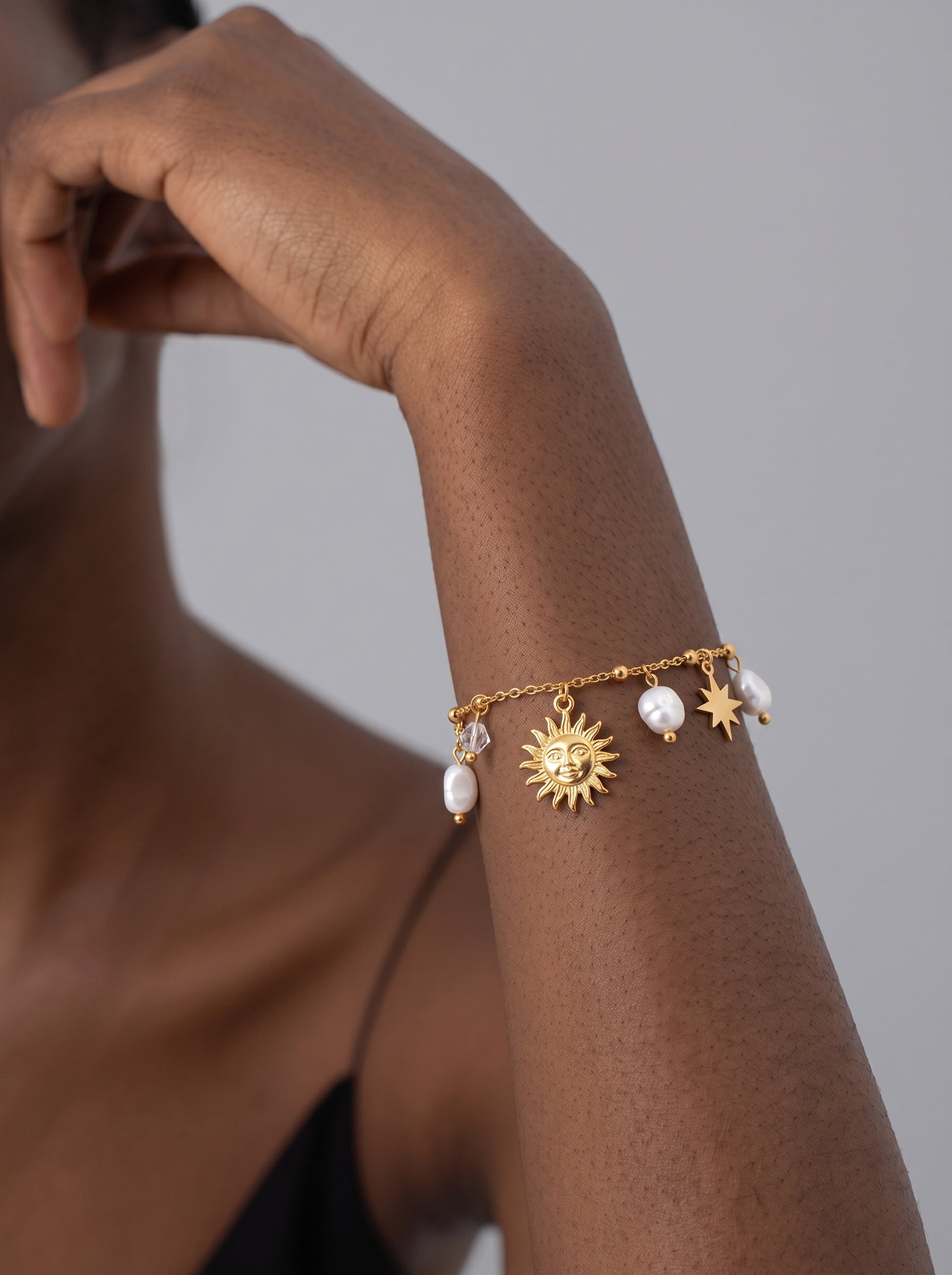 A gold-tone chain bracelet featuring a sun charm, a star charm, baroque freshwater pearls, and faceted beads, worn on a model's arm. @model