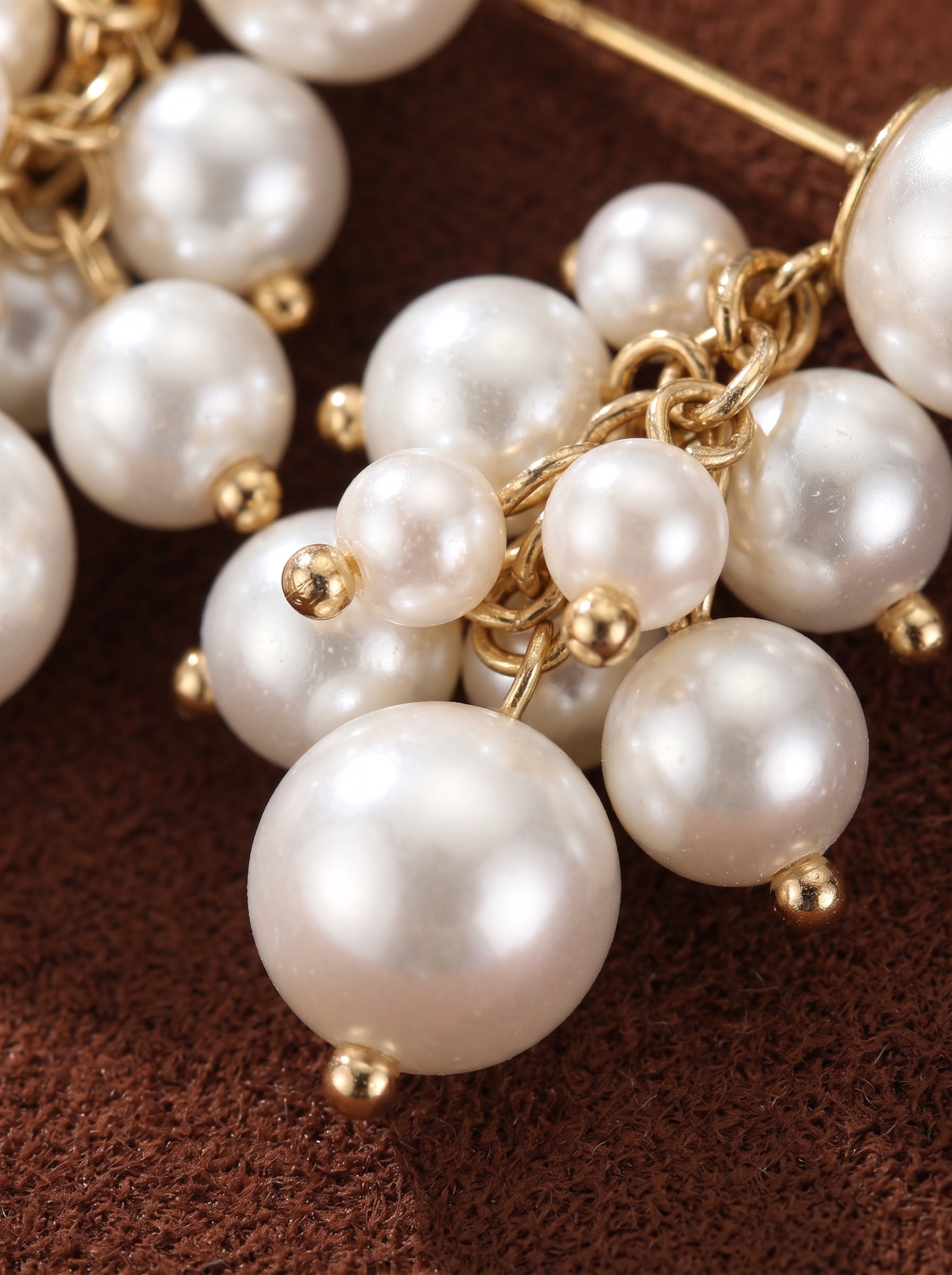 Gros plan des boucles d'oreilles pendantes en grappe Glow Orb présentant une cascade de perles blanches d'eau douce de tailles variées, reliées par de délicates chaînes dorées, contre un fond texturé marron. @pearl