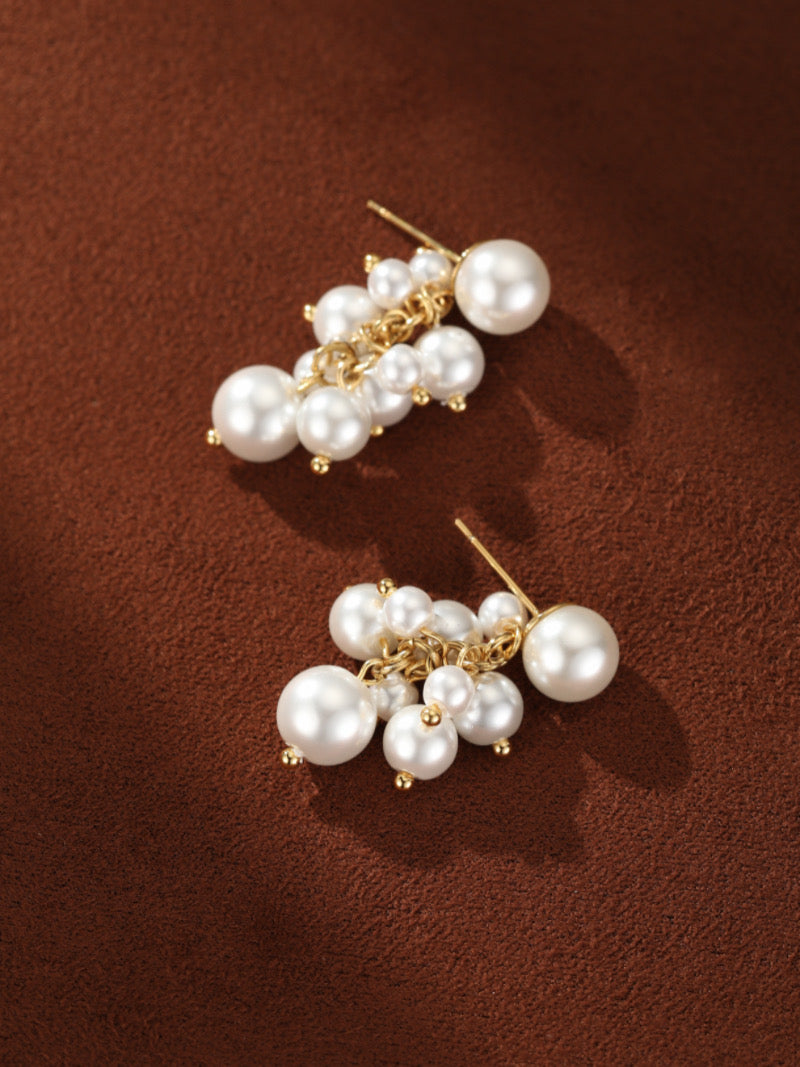 Boucles d'oreilles Glow Orb présentant une puce en grande perle d'eau douce et une cascade de plus petites perles blanches d'eau douce suspendues à une chaîne ton or, contre un fond texturé marron.