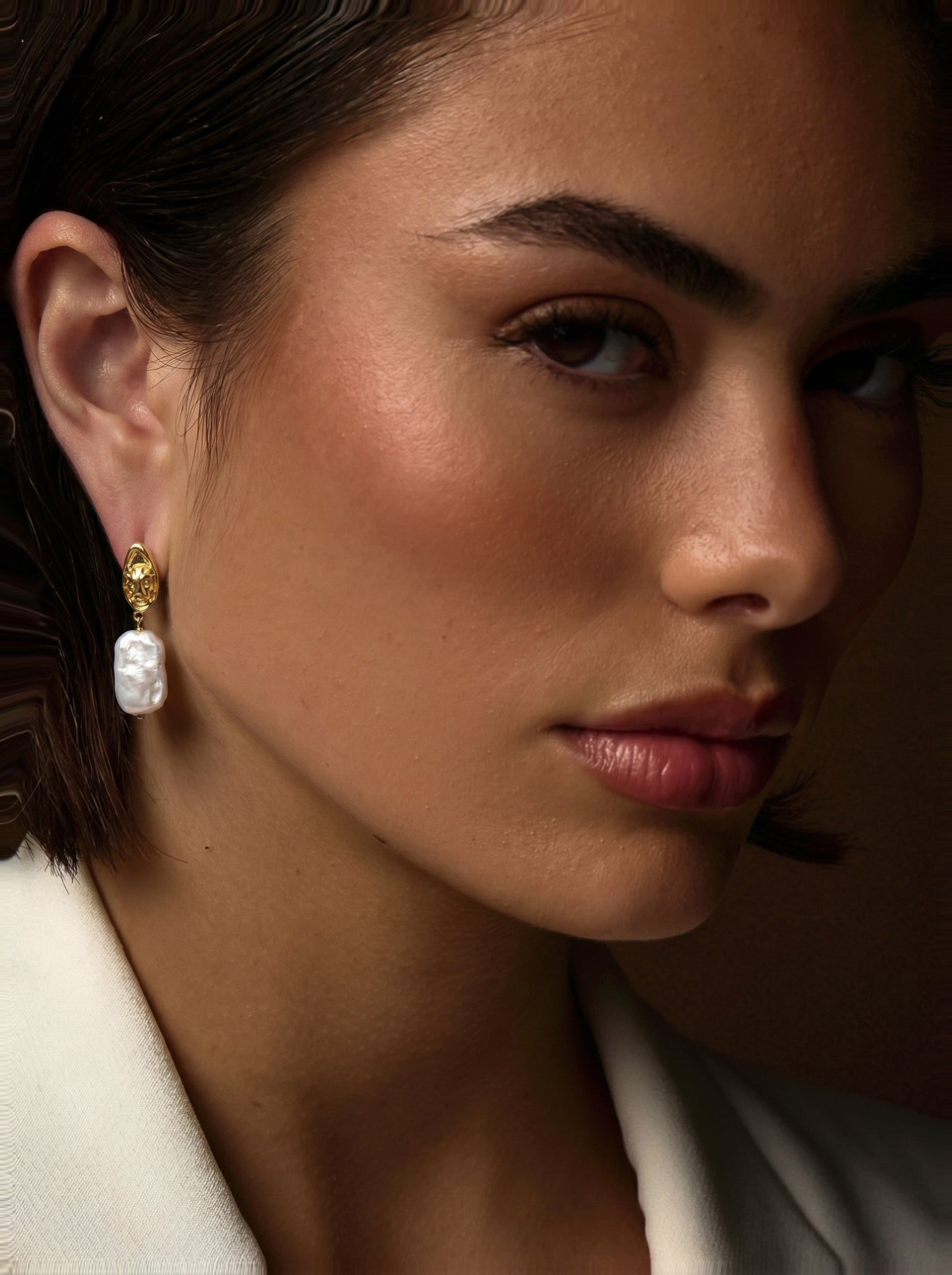 Close-up of a model wearing Glyph Freshwater Pearl Visage Drop Stud Earrings, featuring a gold-toned stud with an embossed visage and a large, irregularly shaped white freshwater pearl drop. @model