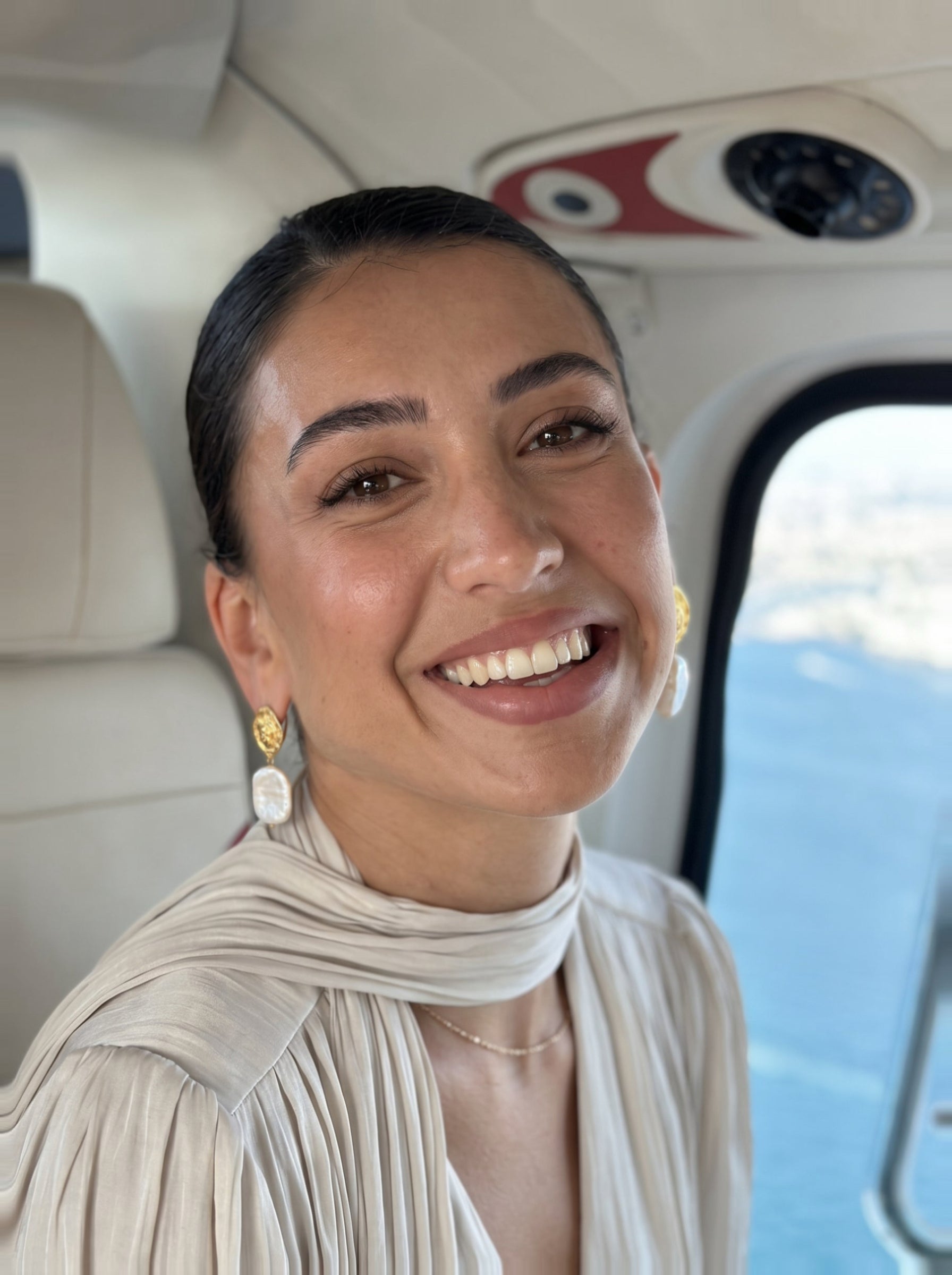A smiling woman wearing Glyph Freshwater Pearl Visage Drop Stud Earrings and a cream-colored outfit, sitting inside a helicopter with a view of the ocean and a ship below. @lifestyle #lifestyle