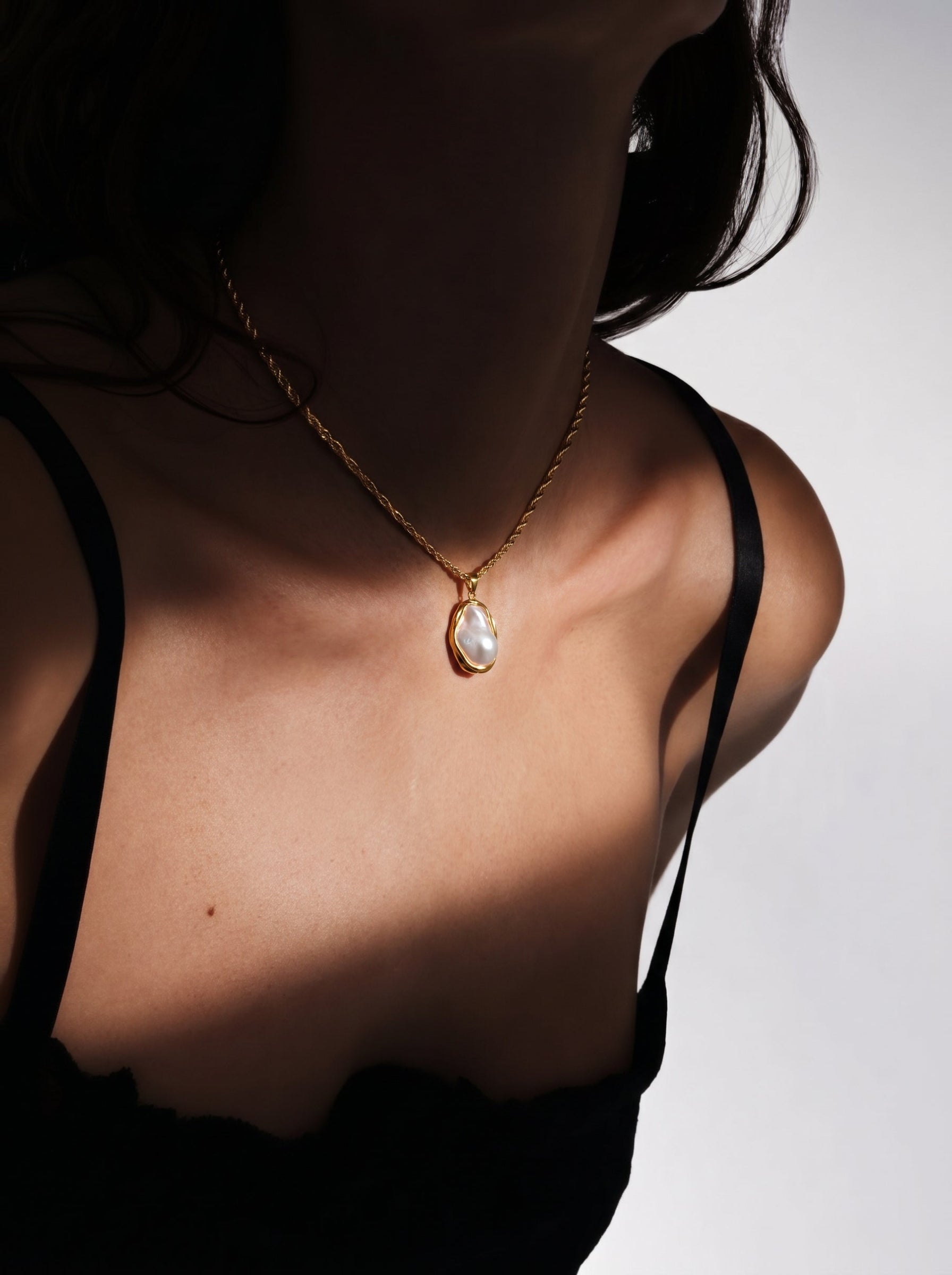 Close-up of a woman wearing a chunky gold chain necklace with a large, irregular white freshwater pearl pendant. The pearl has a natural luster and is set in a fluid gold-toned frame. The necklace is worn over a dark top. @model
