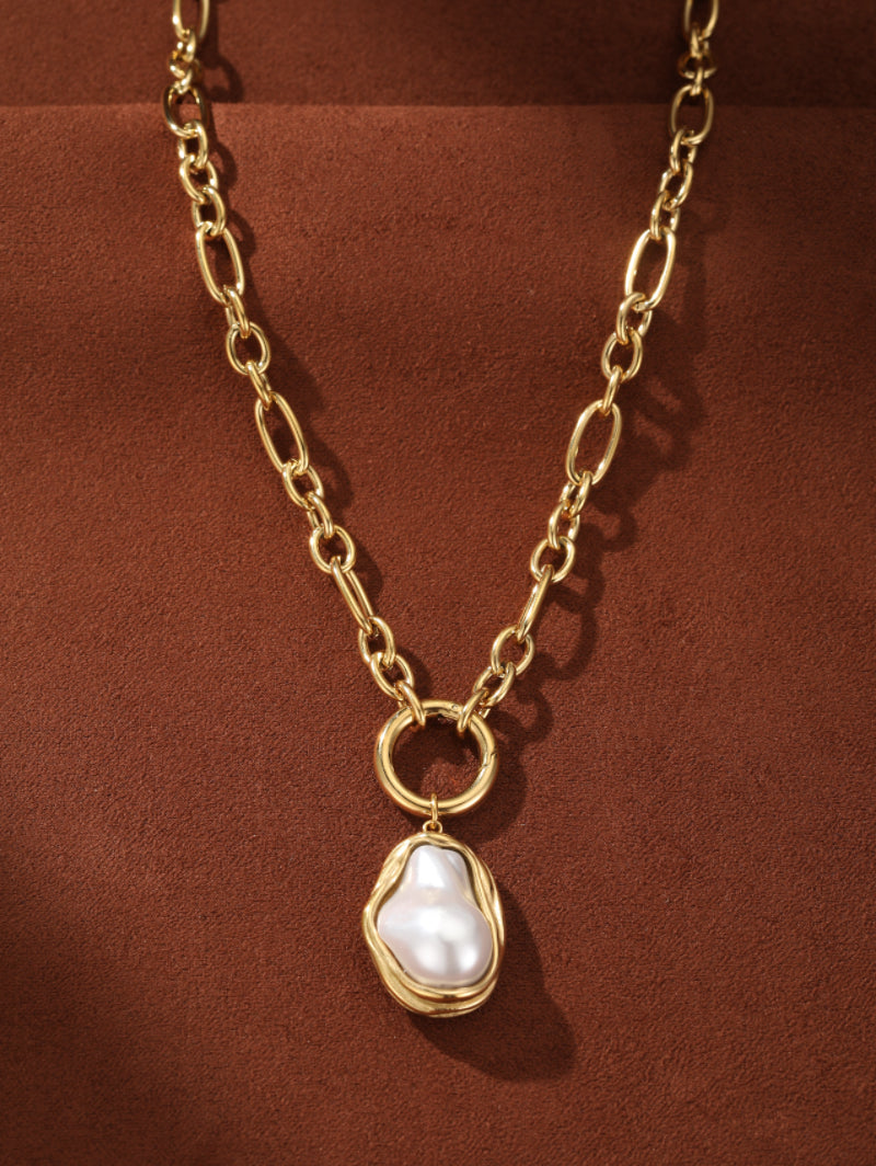 A gold-toned chunky chain necklace features a large, irregular white freshwater pearl pendant with natural luster, set in a fluid gold-toned bezel against a textured brown background.