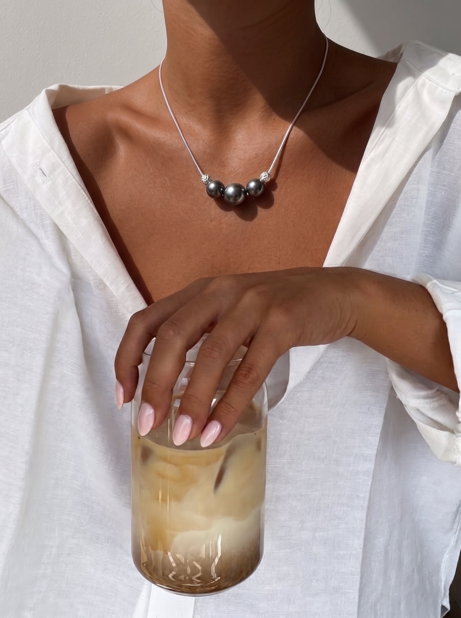 A woman in a white linen shirt holds a glass of iced coffee, wearing a Grit Tahitian Pearl Pendant Necklace with three dark, iridescent pearls separated by textured silver beads on a white cord. @lifestyle