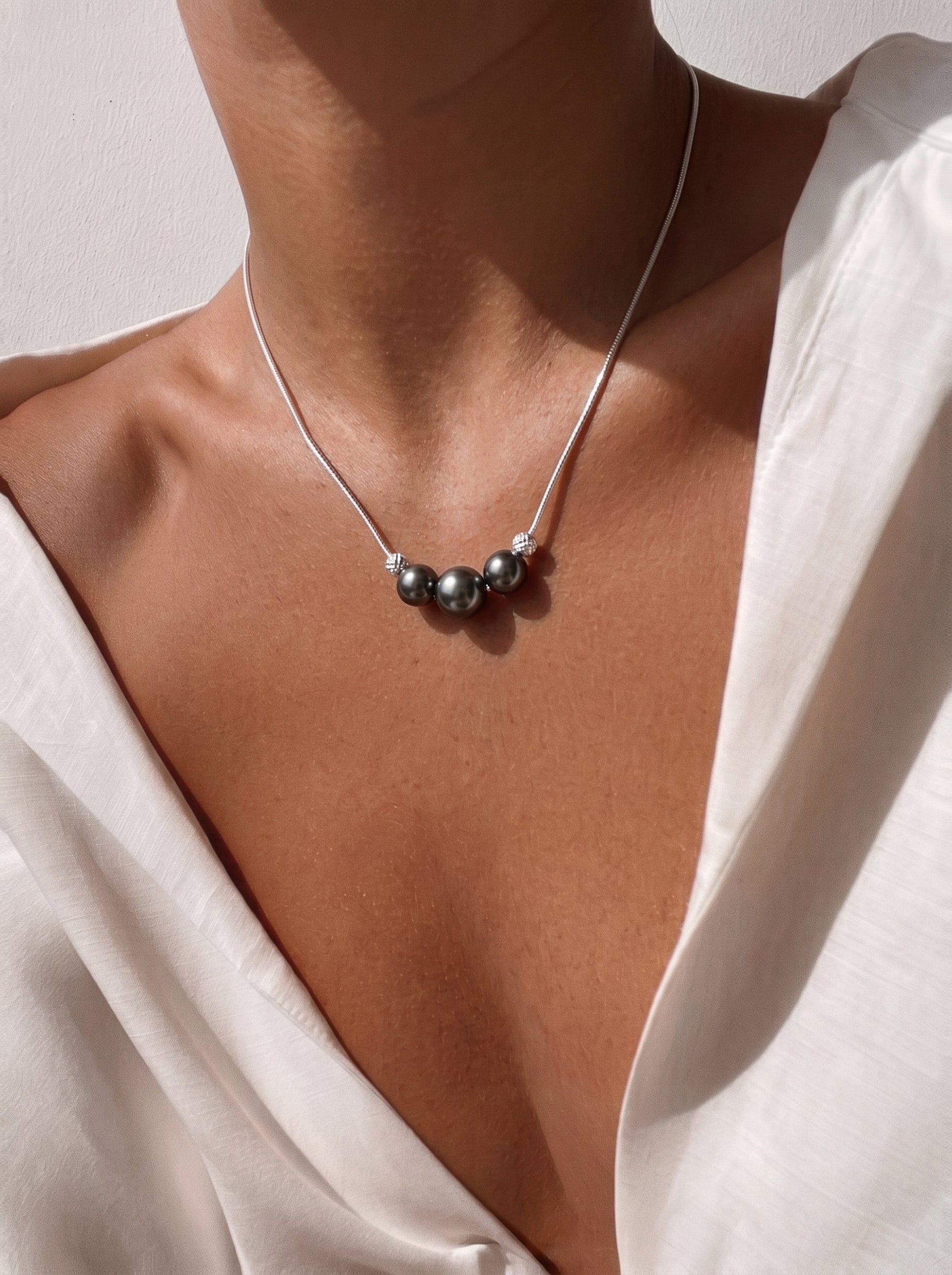 Une femme portant une chemise blanche avec un collier à chaîne serpent argentée présentant trois sombres et lustrées perles de Tahiti séparées par des perles argentées texturées. @model