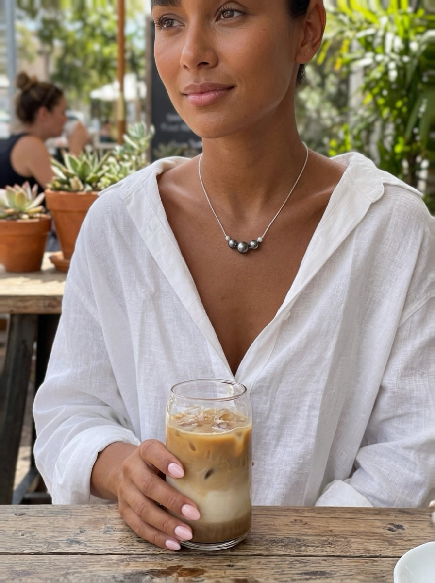 Une femme en chemise en lin blanc tient un verre de café glacé, portant un collier pendentif Grit en perle de Tahiti avec trois perles sombres et irisées séparées par des perles argentées texturées sur un cordon blanc. @lifestyle