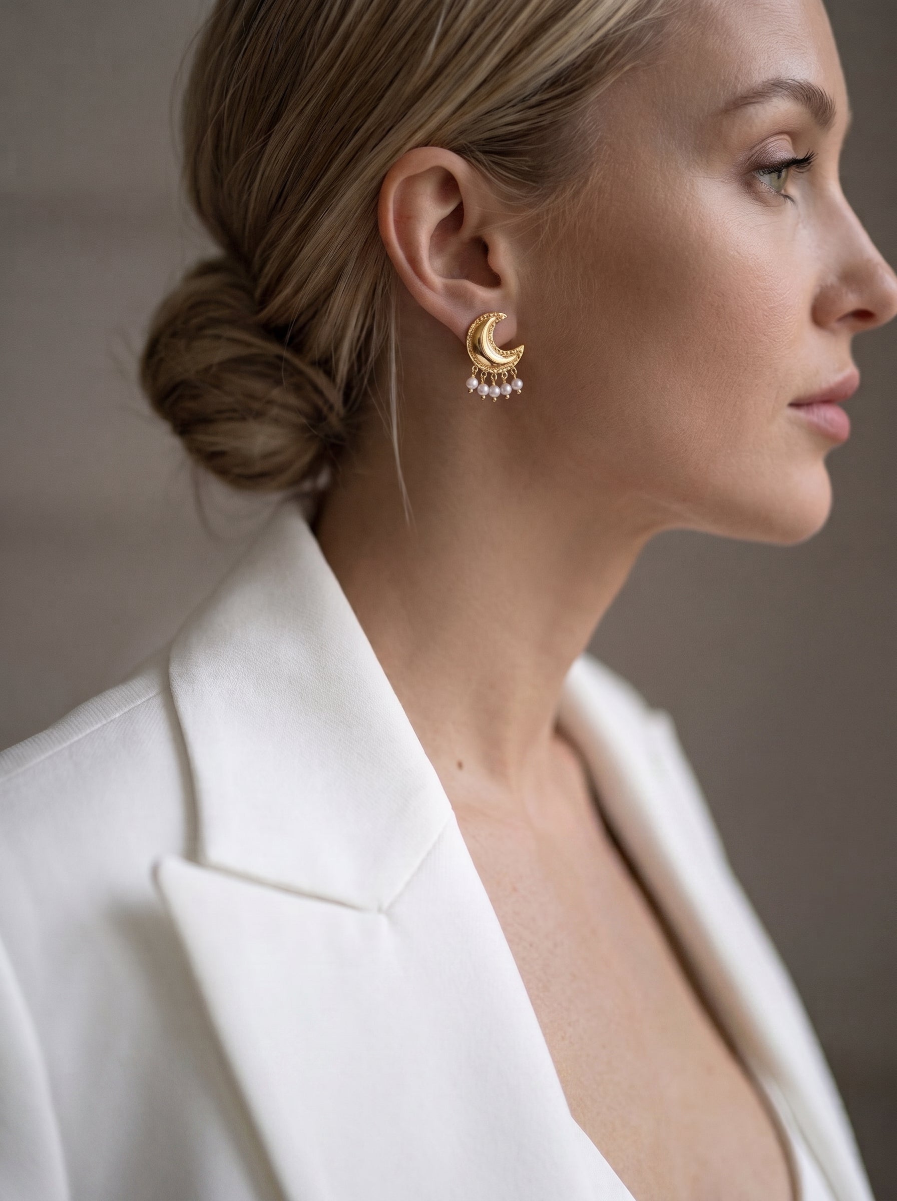 Close-up of a woman wearing Halcyon gold-toned crescent moon drop earrings adorned with five small freshwater pearls, worn with a white blazer. @model