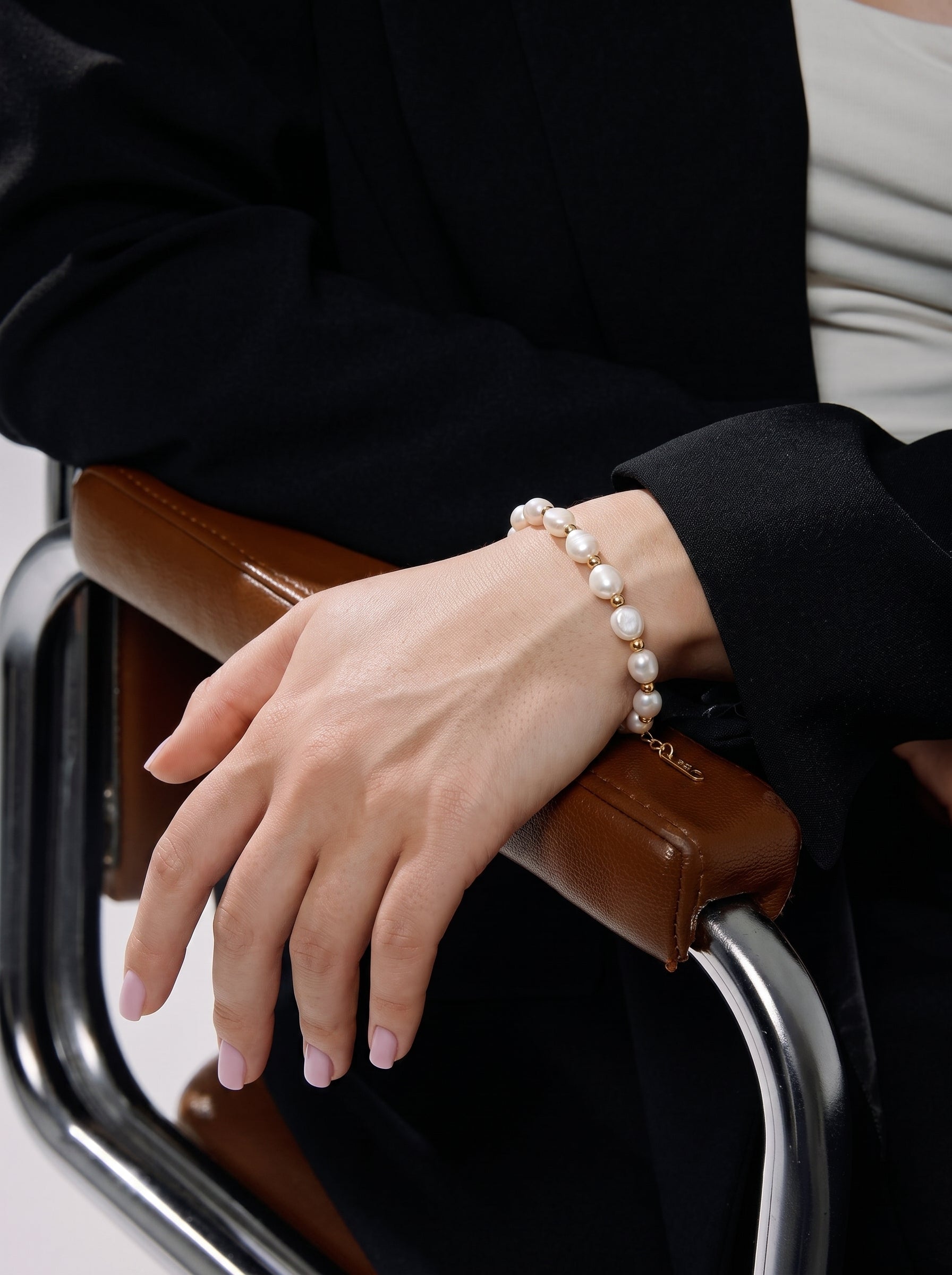 A woman's wrist is adorned with the Inara Freshwater Pearl Beaded Bracelet, featuring lustrous 8mm Baroque pearls interspersed with gold-toned accents, worn with a black blazer and light-colored top. @model