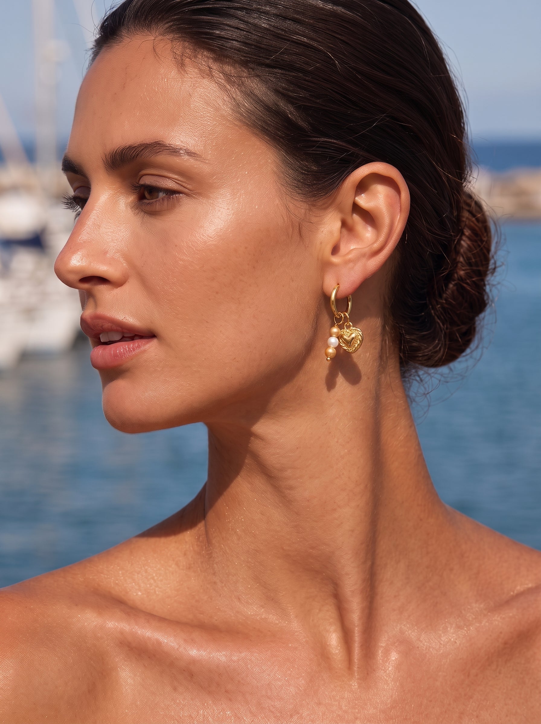Close-up of a woman wearing gold hoop earrings with a heart charm and freshwater pearls, with a blurred background of boats and the sea. @lifestyle