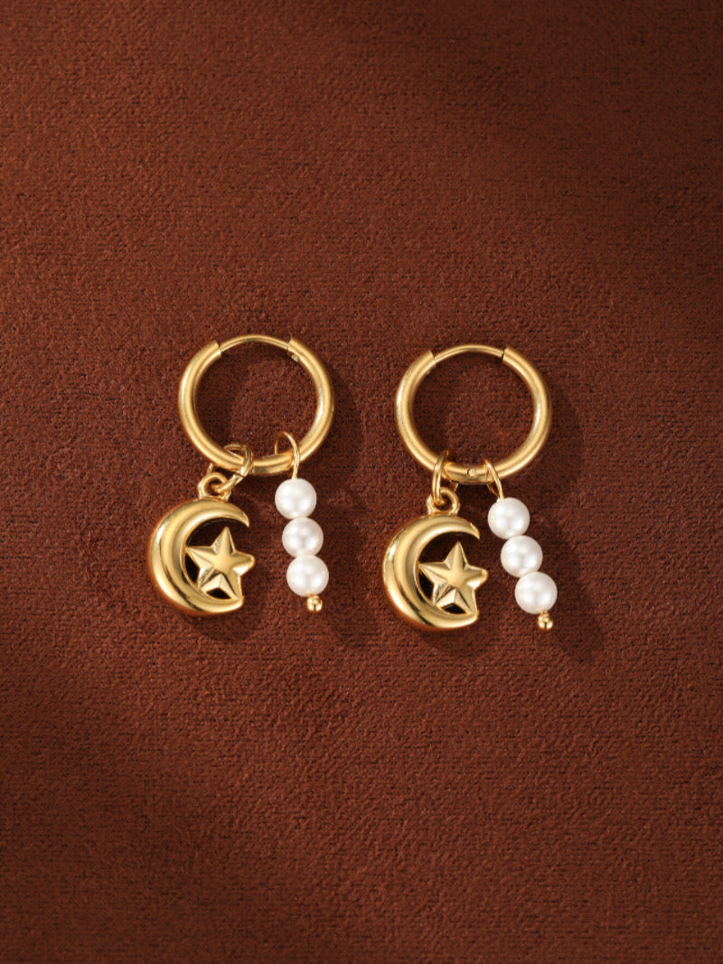 Gold hoop earrings with crescent moon and star charms adorned with a small cluster of freshwater pearls on a brown background.