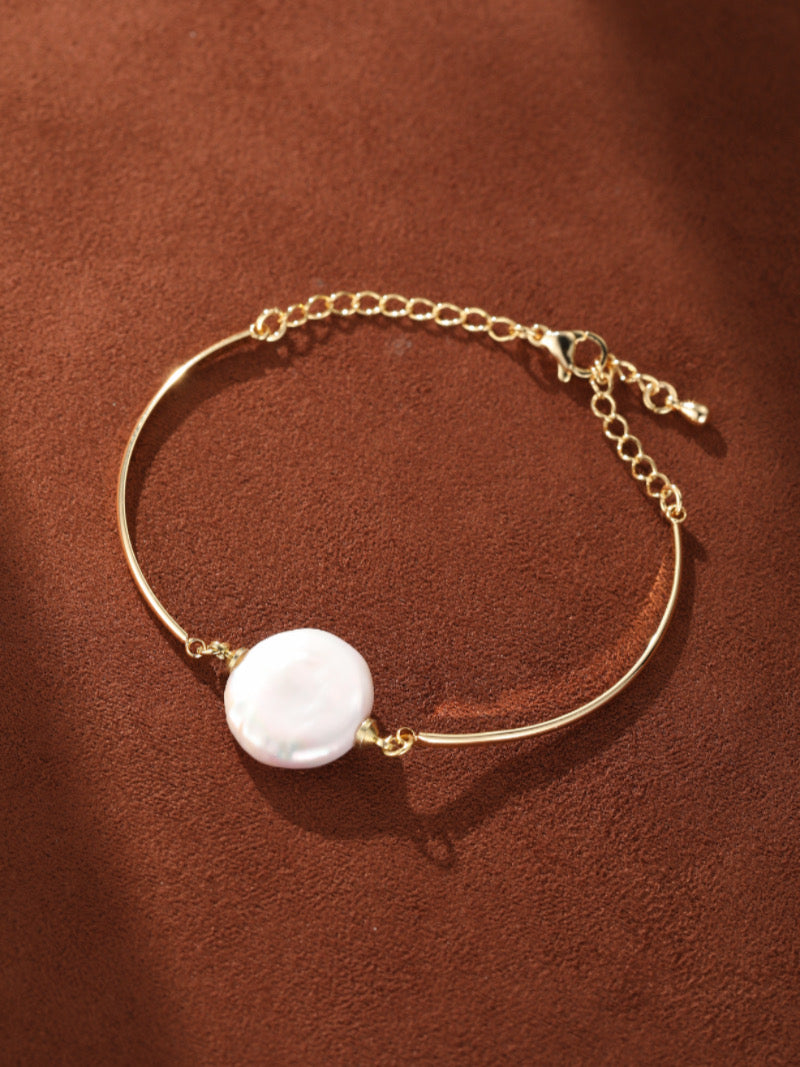Gold-toned adjustable chain bracelet featuring a single, flat, white baroque freshwater pearl suspended centrally. The pearl has a unique, irregular shape, offering a modern and organic aesthetic. The background is a textured brown surface.