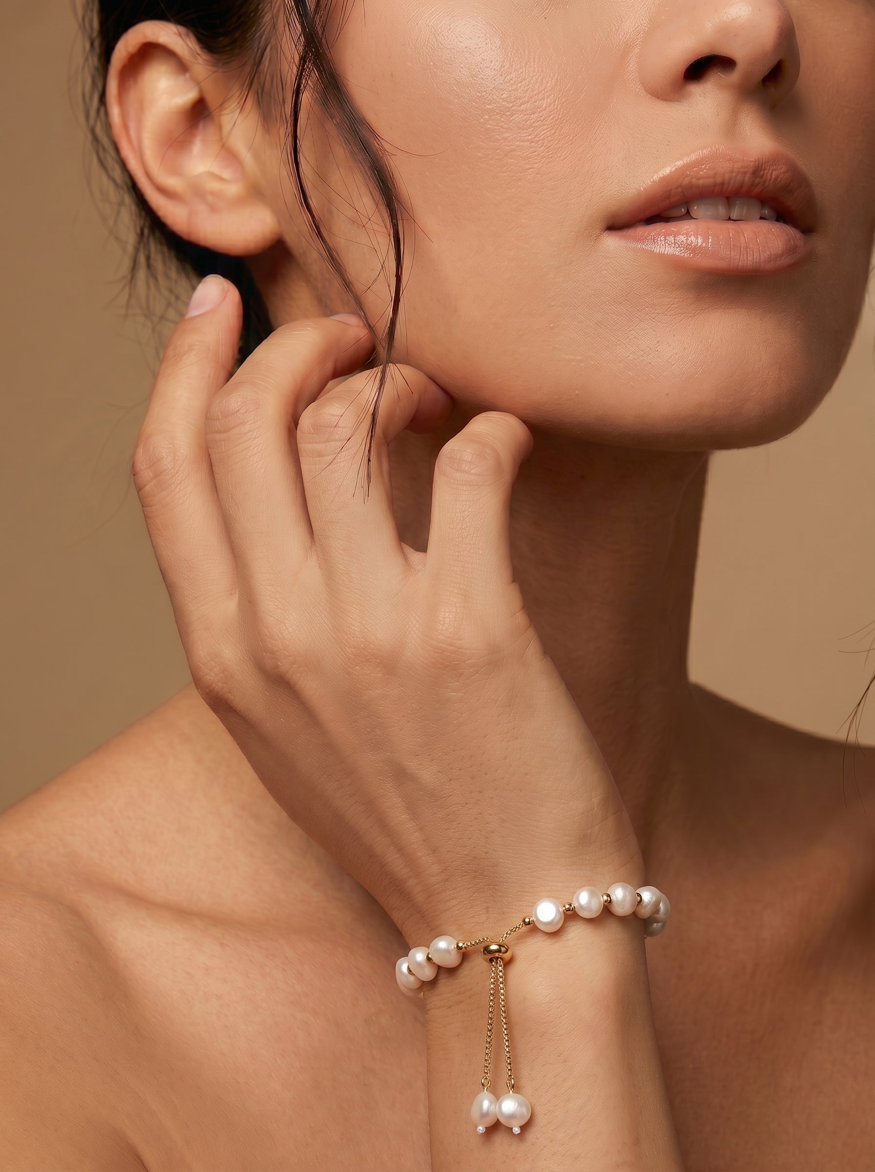 Close-up of a woman's wrist wearing a Kinetic Strand bracelet with 8mm white freshwater pearls and gold-toned beads, featuring an adjustable slider closure. The bracelet has a delicate chain with two small pearls dangling at the end. @model