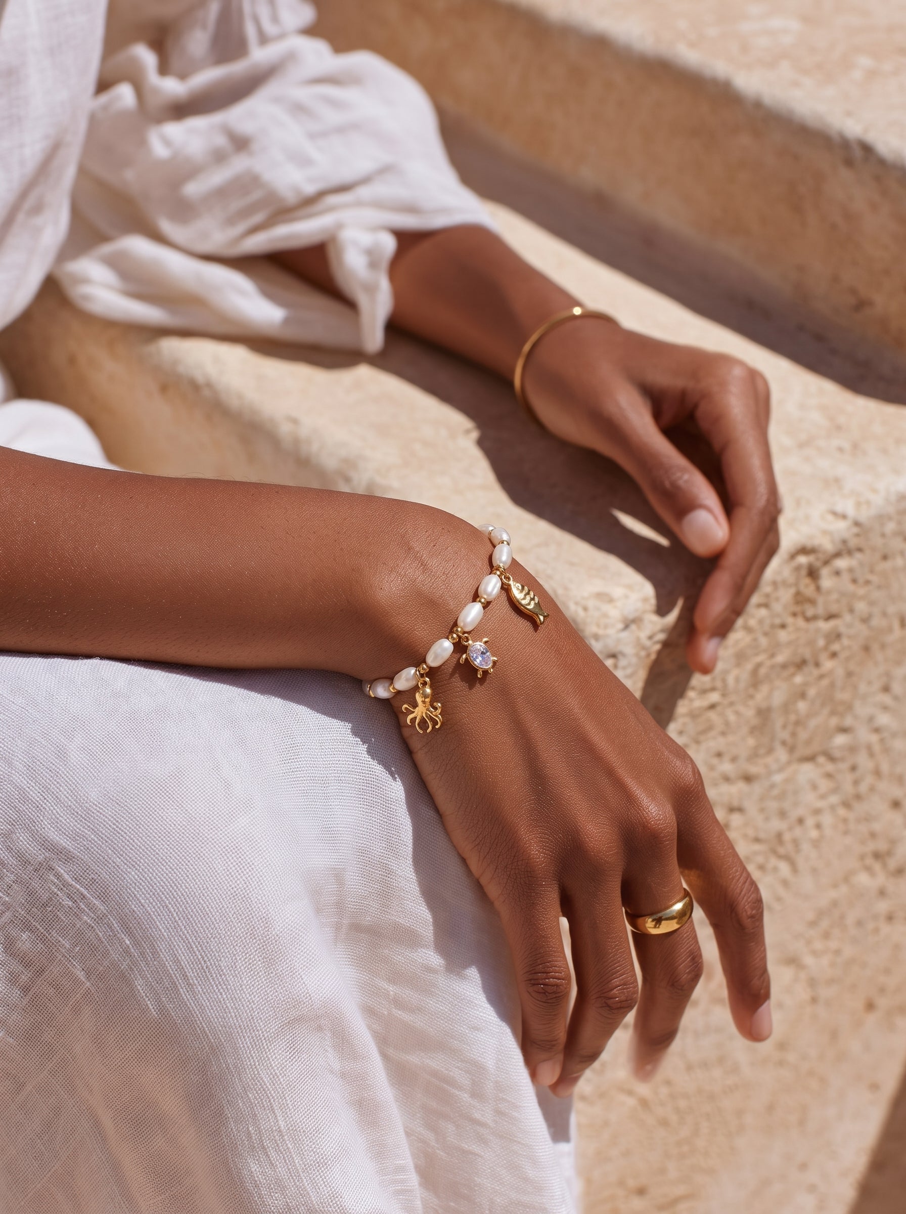 Close-up of a woman's arm wearing a Lagoon Freshwater Pearl Oceanic Charms Beaded Bracelet with gold octopus, fish, and crystal charms, styled with a white linen dress and gold ring. @model