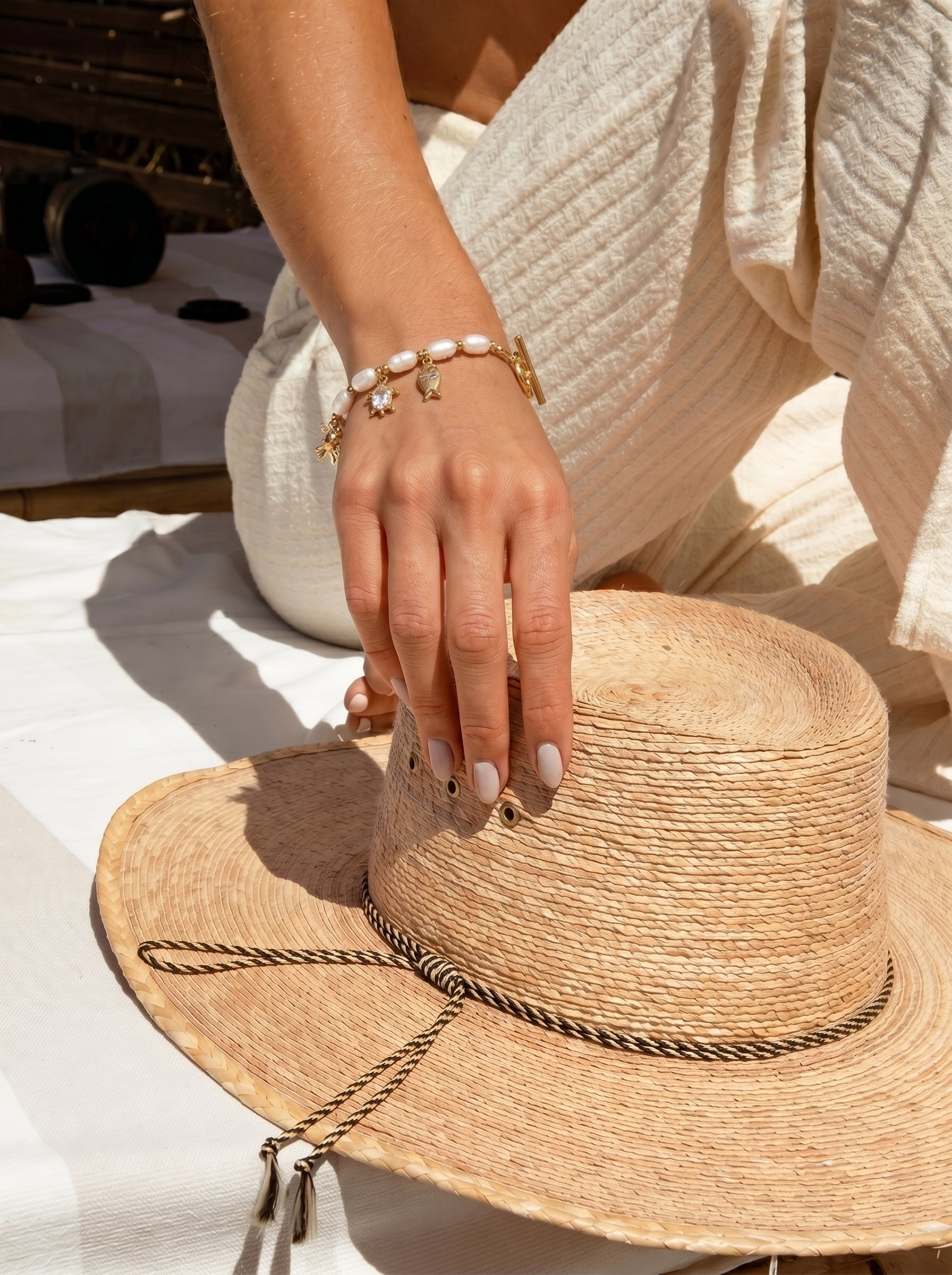 A woman's hand wearing a Lagoon Freshwater Pearl Oceanic Charms Beaded Bracelet, featuring white pearls and gold charms like a starfish and whale, rests on a straw hat. The scene evokes a sunny, relaxed summer holiday vibe. @lifestyle