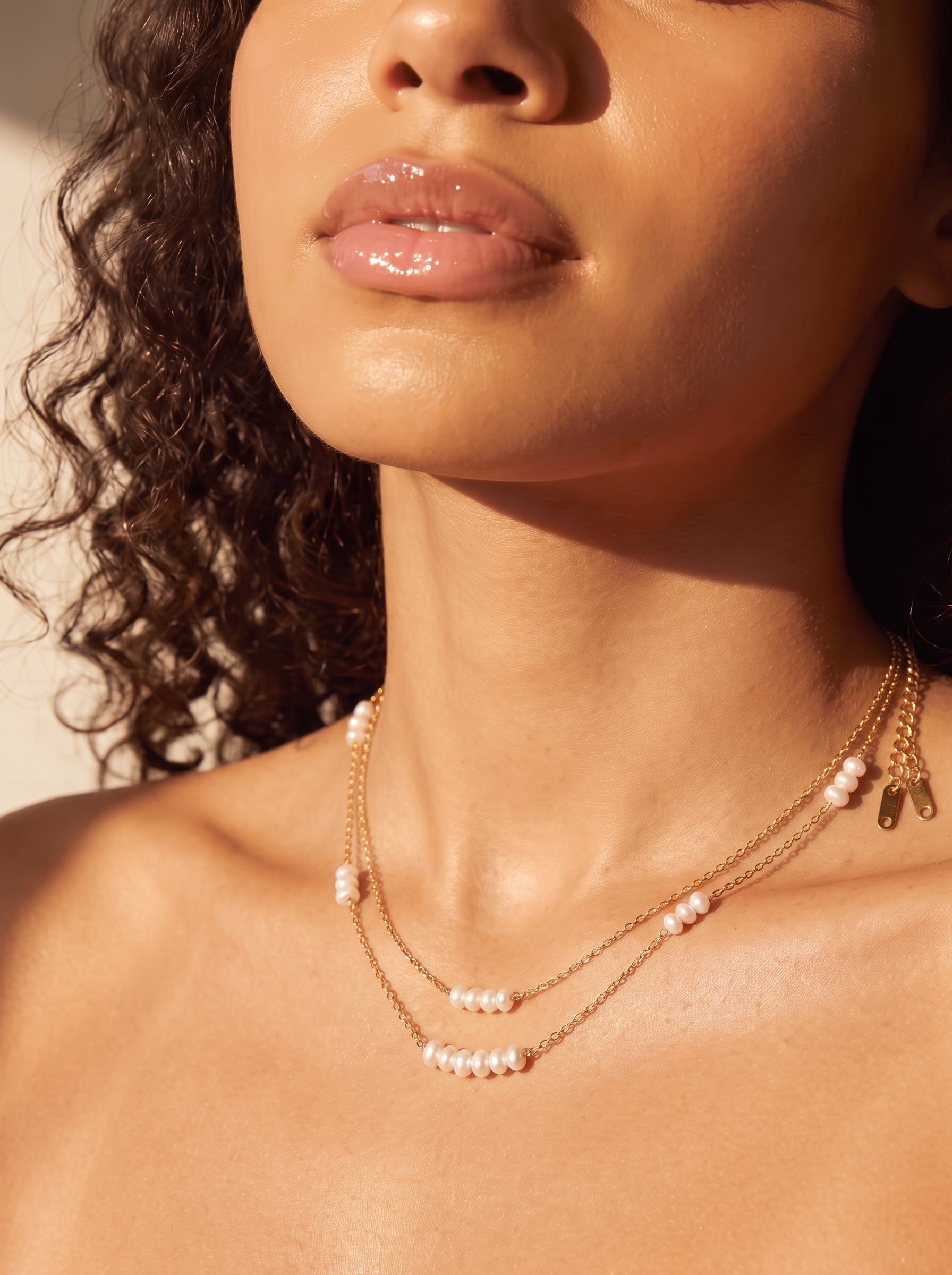 Gros plan du décolleté et des lèvres d'une femme portant un délicat collier superposé doré avec de petites perles graines blanches d'eau douce espacées le long des chaînes. @model