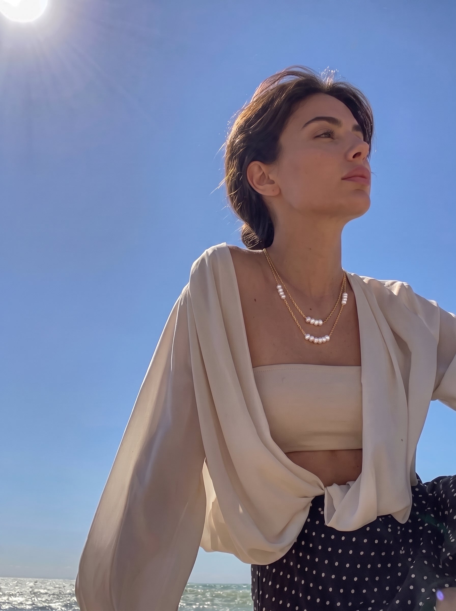 Une femme portant le collier superposé perlé Lithe Thread en perle d'eau douce par une journée ensoleillée au bord de la mer, avec l'océan et un ciel bleu vif en arrière-plan. Le collier présente deux délicates chaînes ton or avec de petites perles graines blanches espacées le long, créant un effet superposé. La femme regarde au loin, incarnant une présence sereine et confiante. @lifestyle