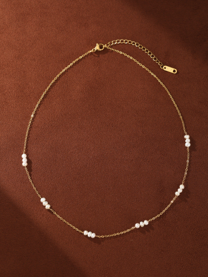 Délicat collier à chaîne ton or présentant de petites perles graines blanches d'eau douce espacées uniformément le long d'un seul rang, présenté contre un fond marron.