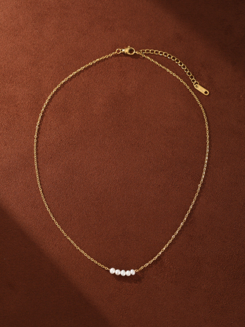 Un délicat collier à chaîne ton or avec sept petites perles graines blanches d'eau douce espacées uniformément le long du centre, sur fond marron chaleureux.