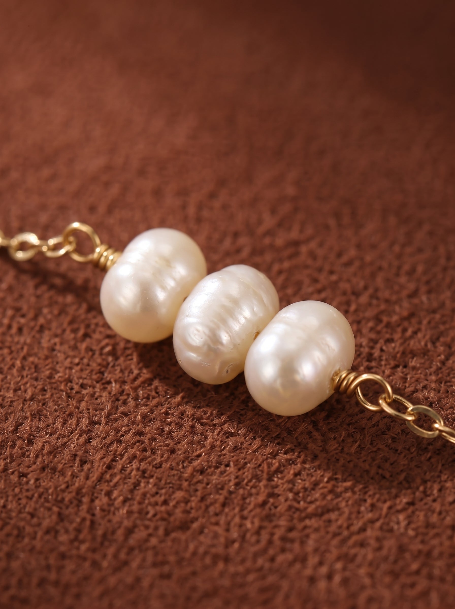 Gros plan de trois perles graines blanches d'eau douce sur une délicate chaîne ton or, mettant en valeur leur éclat et leur texture sur fond texturé marron. @pearl