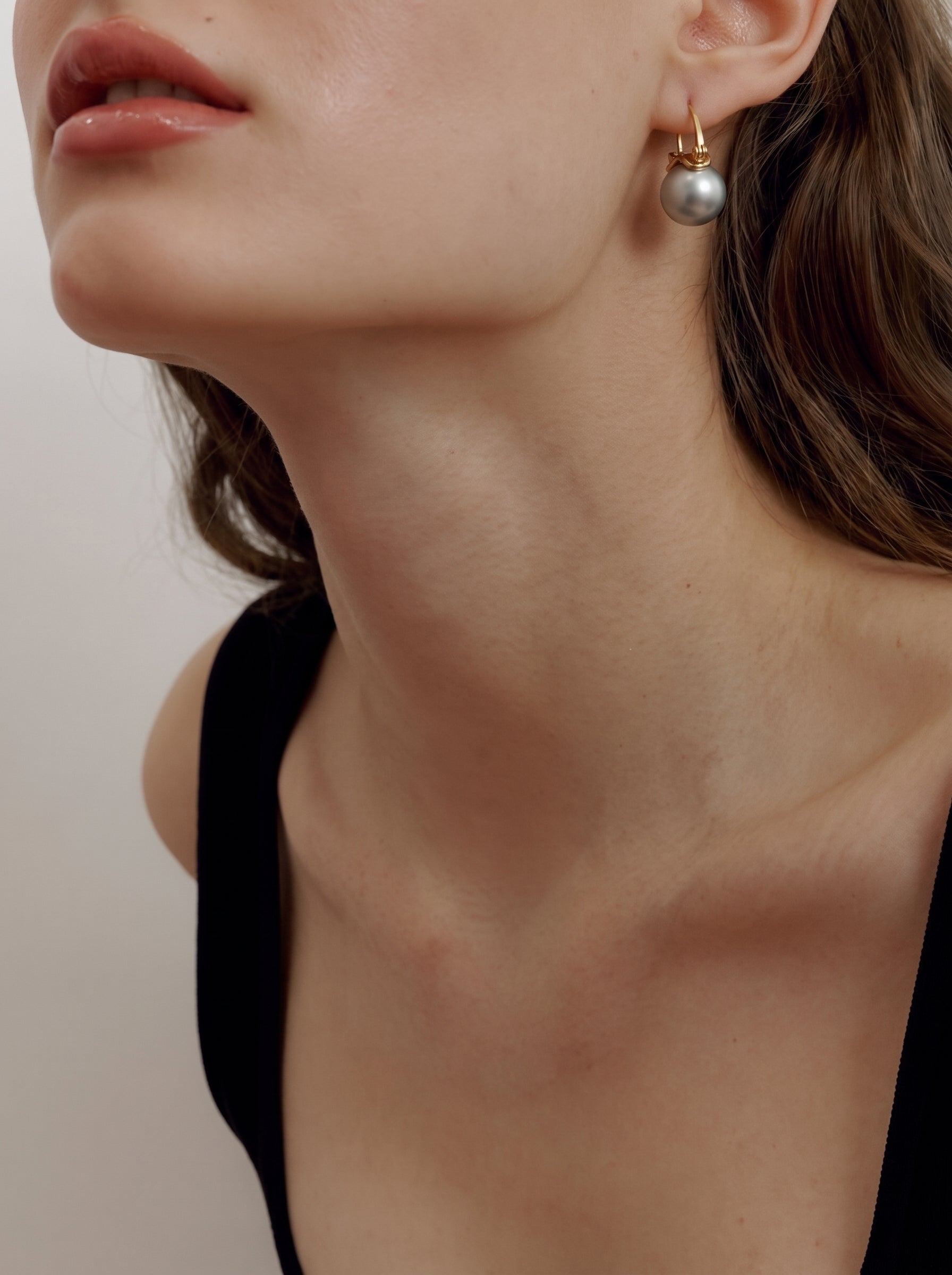 Close-up of a woman wearing Lode Freshwater Pearl Hoop Drop Earrings, featuring a large, round, grey pearl suspended from a gold-toned hoop. @model
