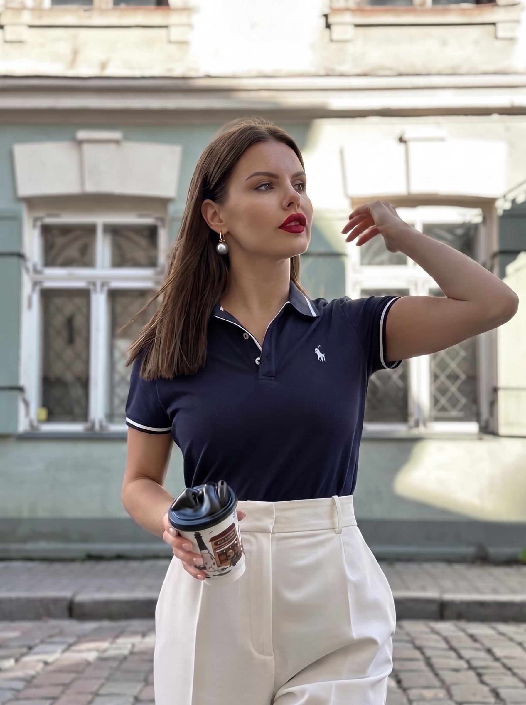 A woman wearing a navy polo shirt and cream-colored pants stands outside, holding a coffee cup and wearing Lode freshwater pearl hoop drop earrings. @lifestyle