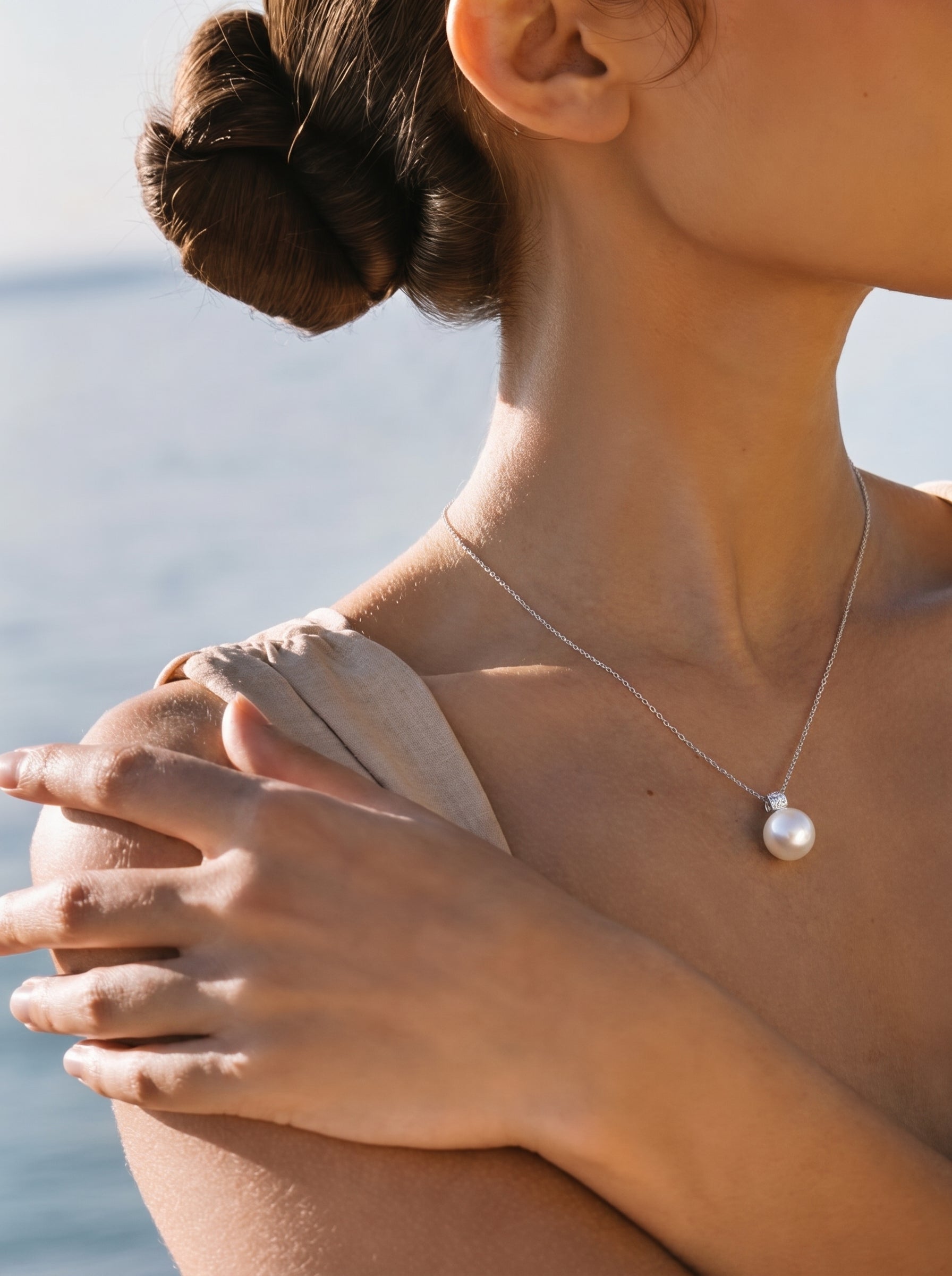 Gros plan d'une femme portant un collier pendentif à chaîne Lumin en perle des mers du Sud avec une grande et lustrée perle blanche et un petit accent diamant contre une toile de fond d'océan bleu doux. @lifestyle