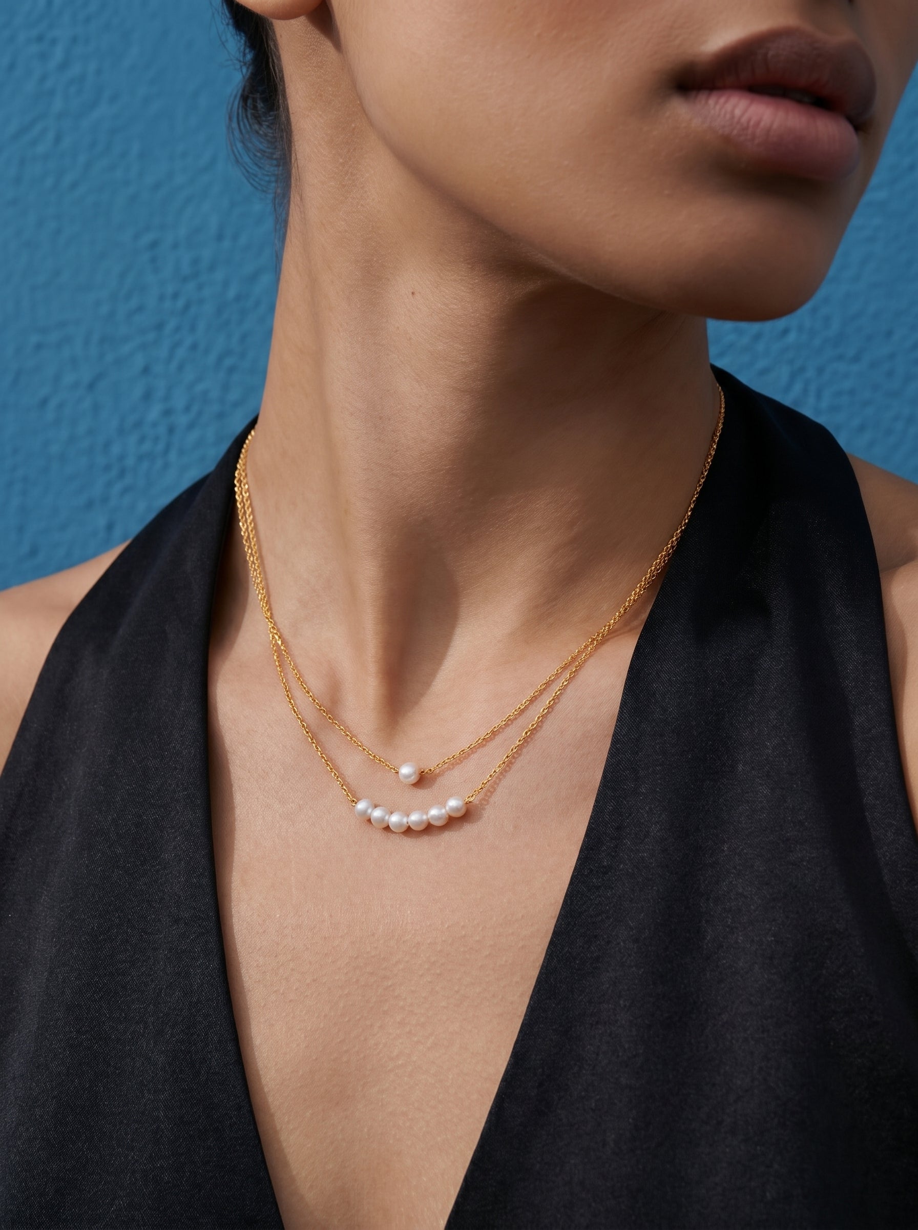 Close-up of a model wearing the Lumina layered chain necklace with small white imitation pearls against a blue background. #model
