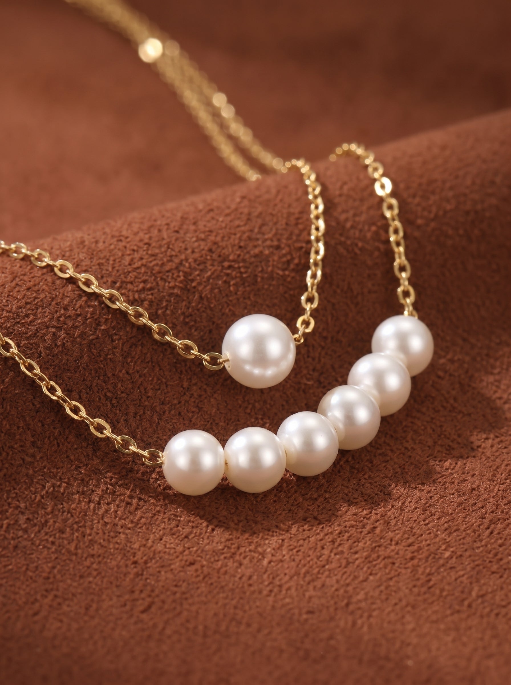 A layered gold chain necklace featuring multiple round white imitation pearls on a textured brown background. The necklace is delicate and elegant, with pearls of varying sizes.