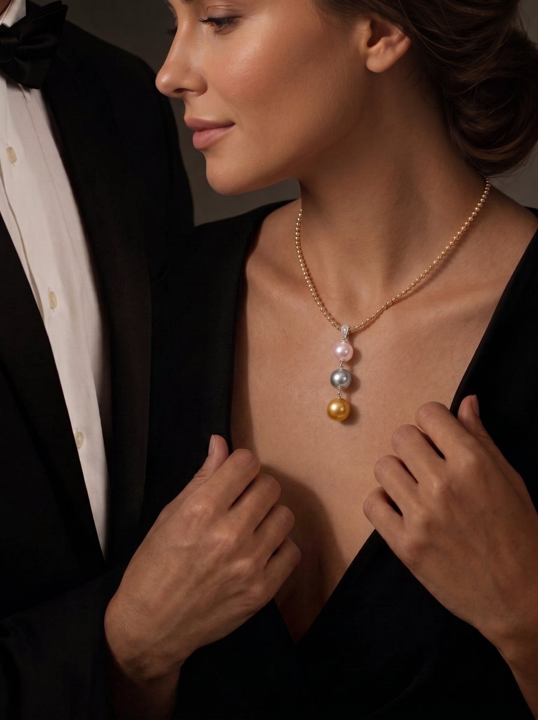 A woman in a black dress and a man in a tuxedo embrace, showcasing the Luminance necklace with three pearls of varying colors: white, gray, and gold, dangling from a long, mixed-tone chain. @lifestyle