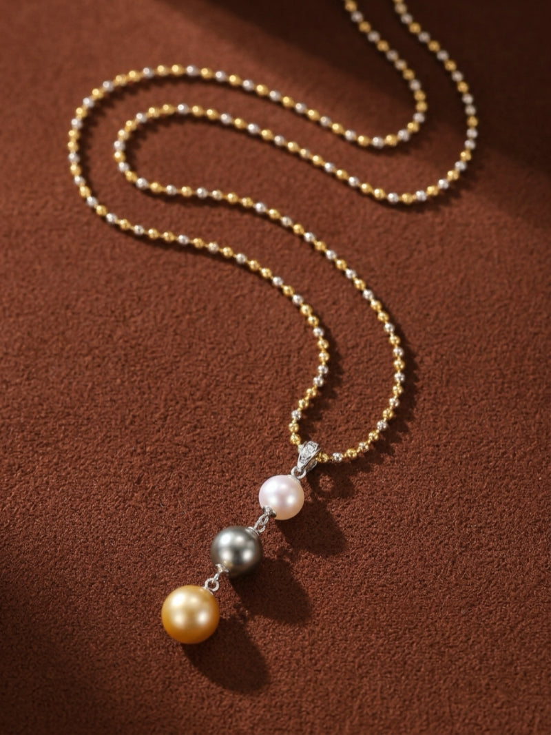 Luminance necklace featuring a long ball chain with alternating gold and silver tones, suspending three round pearls: a white Akoya pearl, a grey Tahitian pearl, and a golden South Sea pearl.