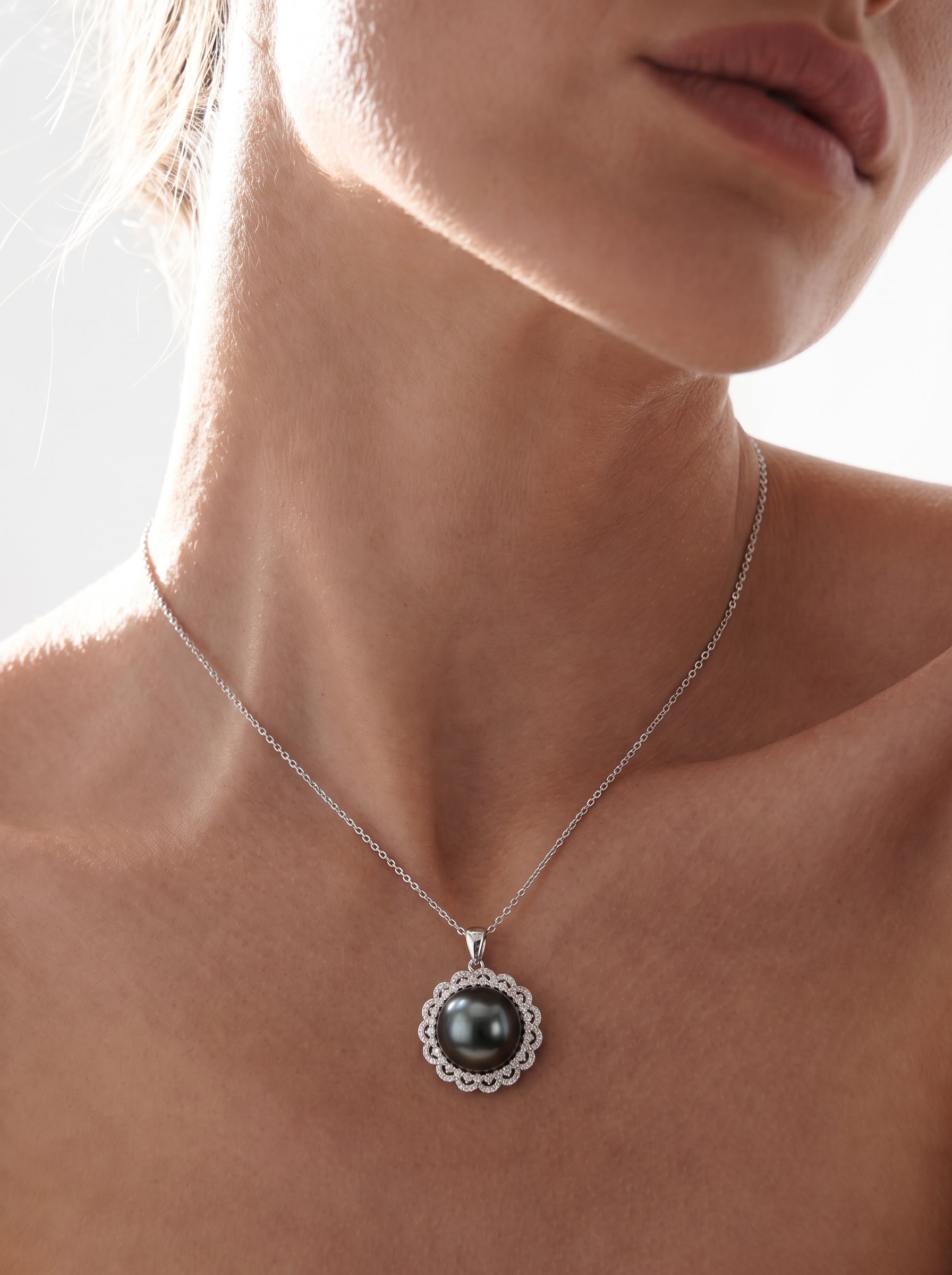 A close-up shot of a woman wearing the Luminary Tahitian Mabe Pearl Sun Pendant Necklace, featuring a large black pearl surrounded by a sunburst halo of shimmering accents on a delicate silver chain. @model