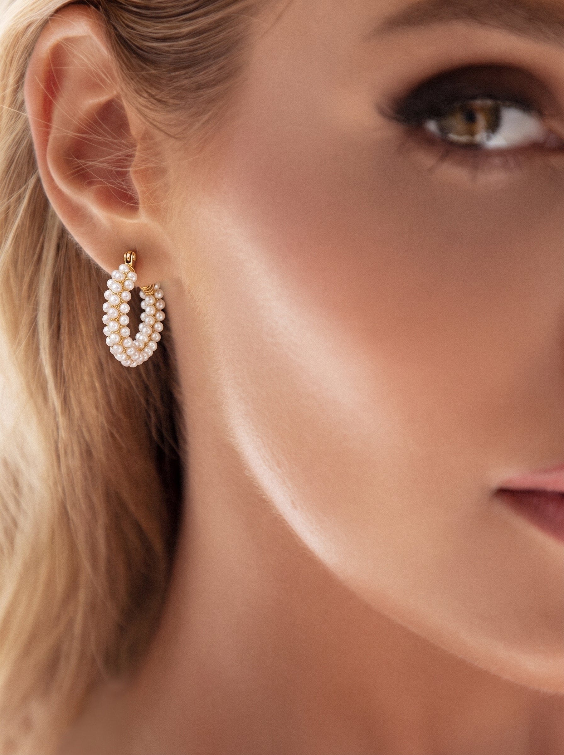 Close-up of a woman's earlobe wearing Luminous Bloom chunky hoop earrings, adorned with multiple rows of small white imitation pearls on a gold-toned base. The earrings are oval-shaped and textured, offering a chic and radiant look. @model