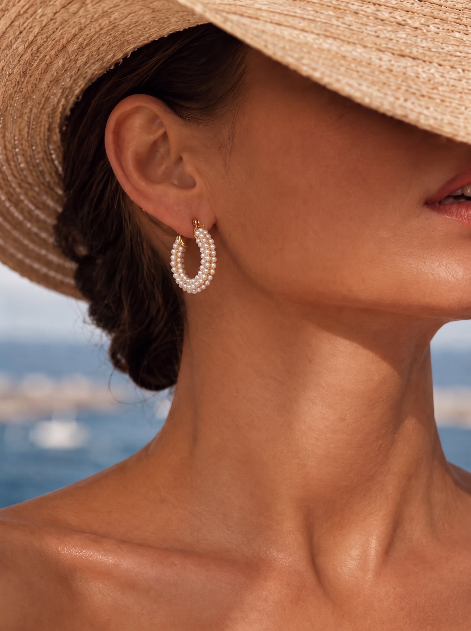 A close-up shot of a woman wearing Luminous Bloom Imitation Pearl Chunky Hoop Earrings, with a straw hat partially obscuring her face and a blurred ocean background. @lifestyle