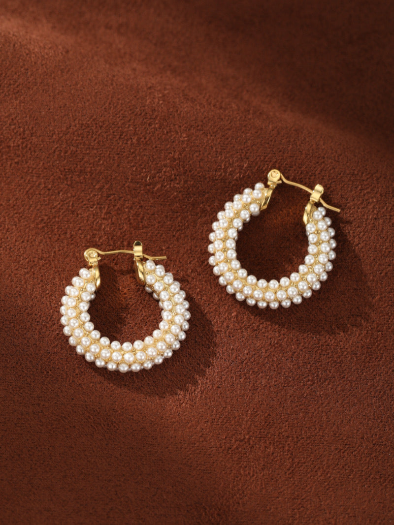 Chunky gold-toned hoop earrings densely adorned with multiple rows of lustrous white imitation pearls on a brown textured background.